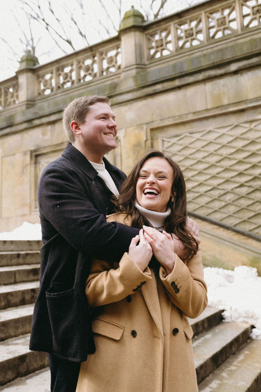Couple laughing and embracing on stone steps during a winter engagement photoshoot.