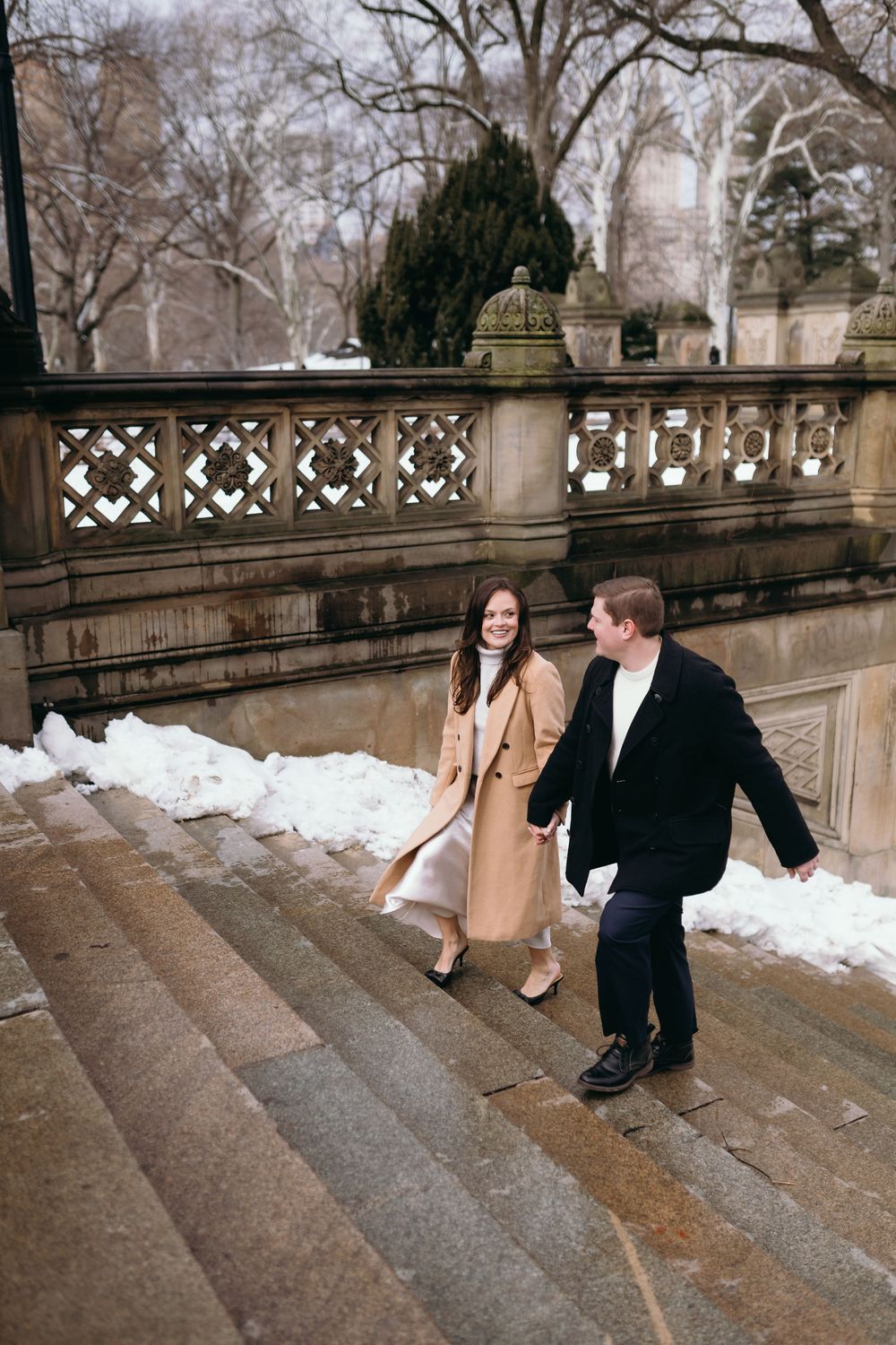 A couple walks down snowy stone steps in a romantic winter scene at a historic location.