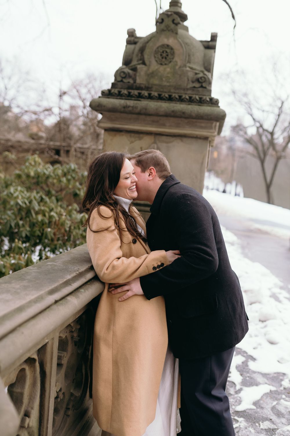 A romantic winter engagement photo of a couple kissing by an ornate stone railing in the snow.