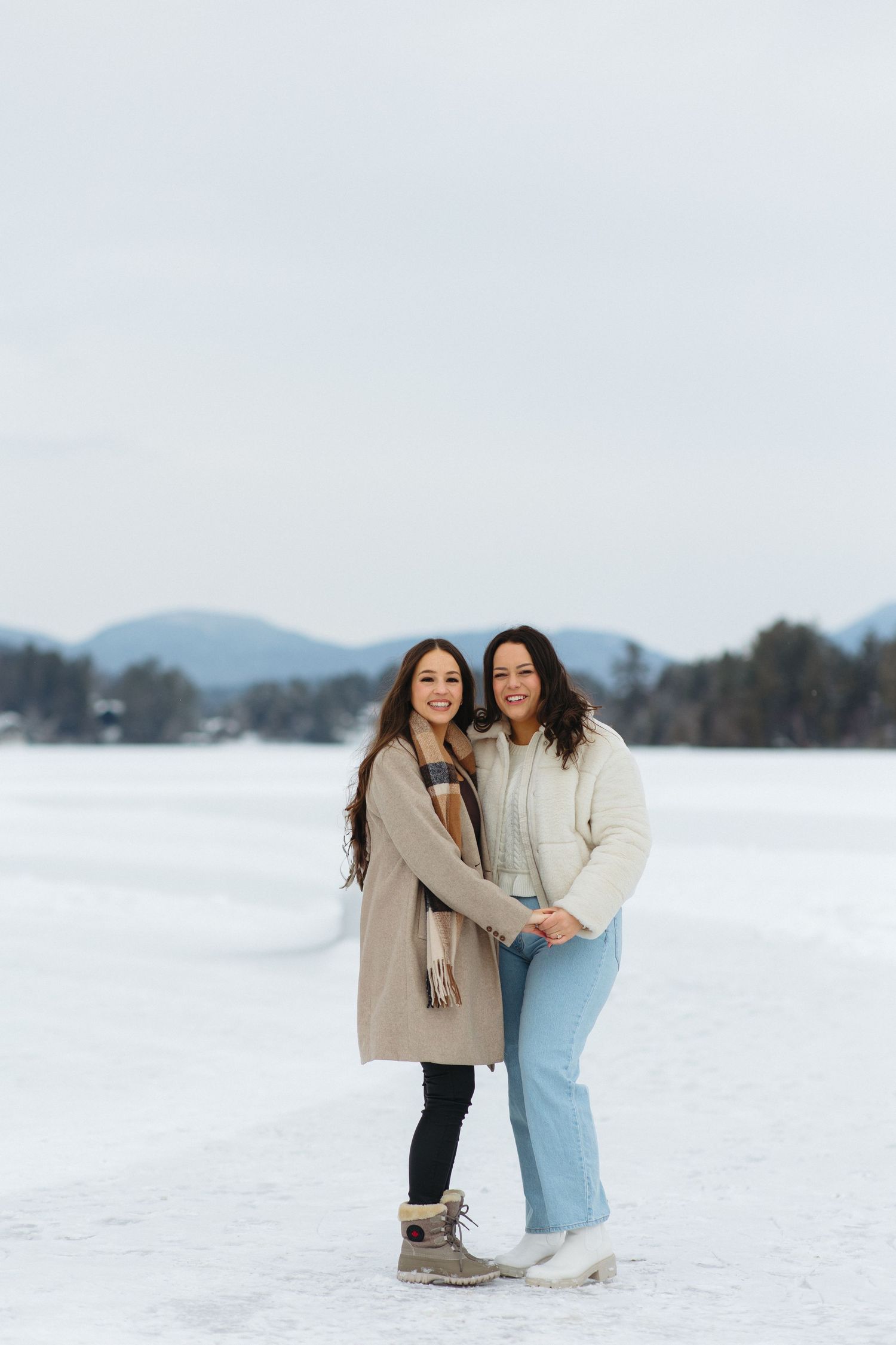 Two people embracing and smiling while standing in a snow-covered field with mountains in the background.