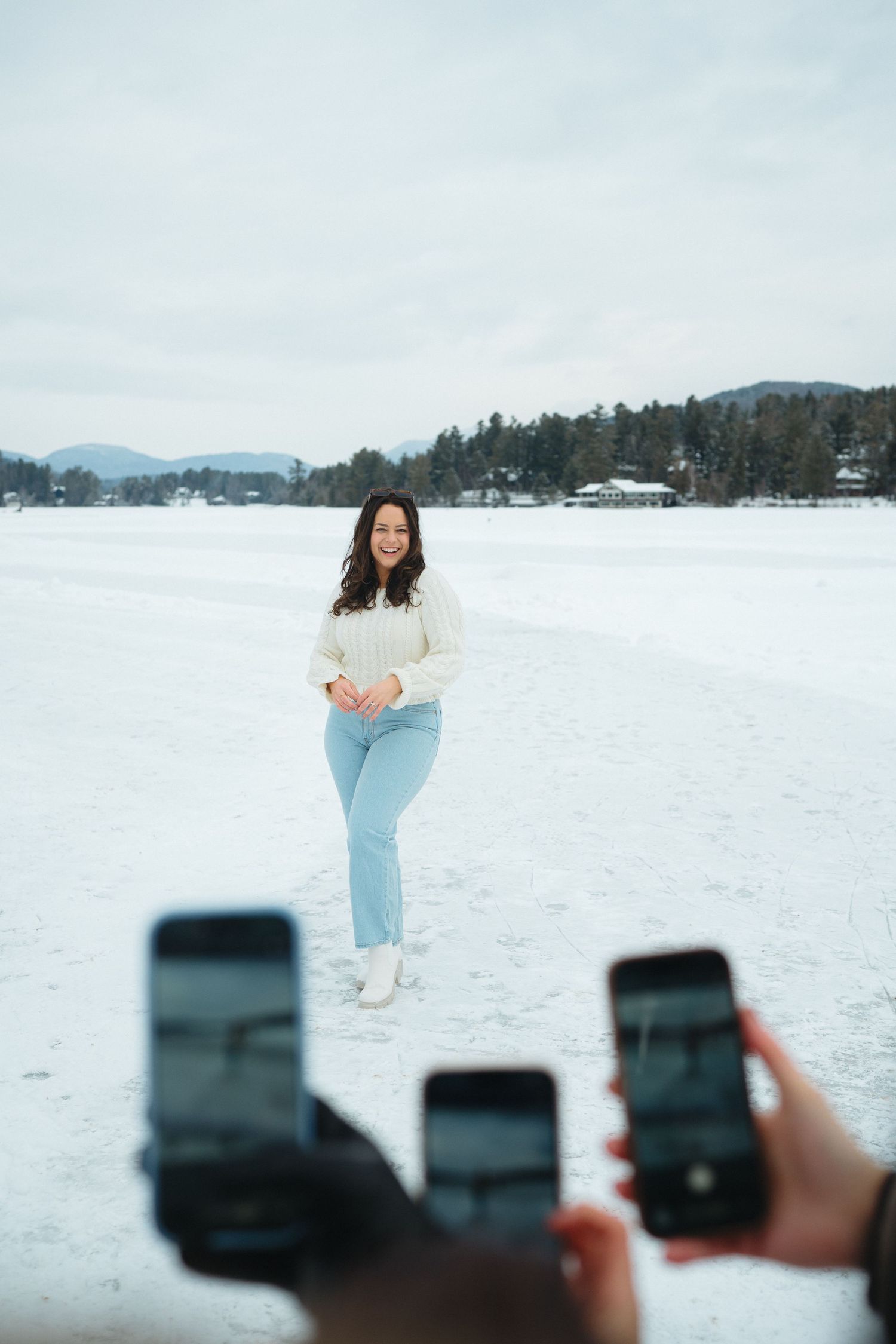 Three phones held up to photograph someone posing in a white sweater on a snowy winter landscape.