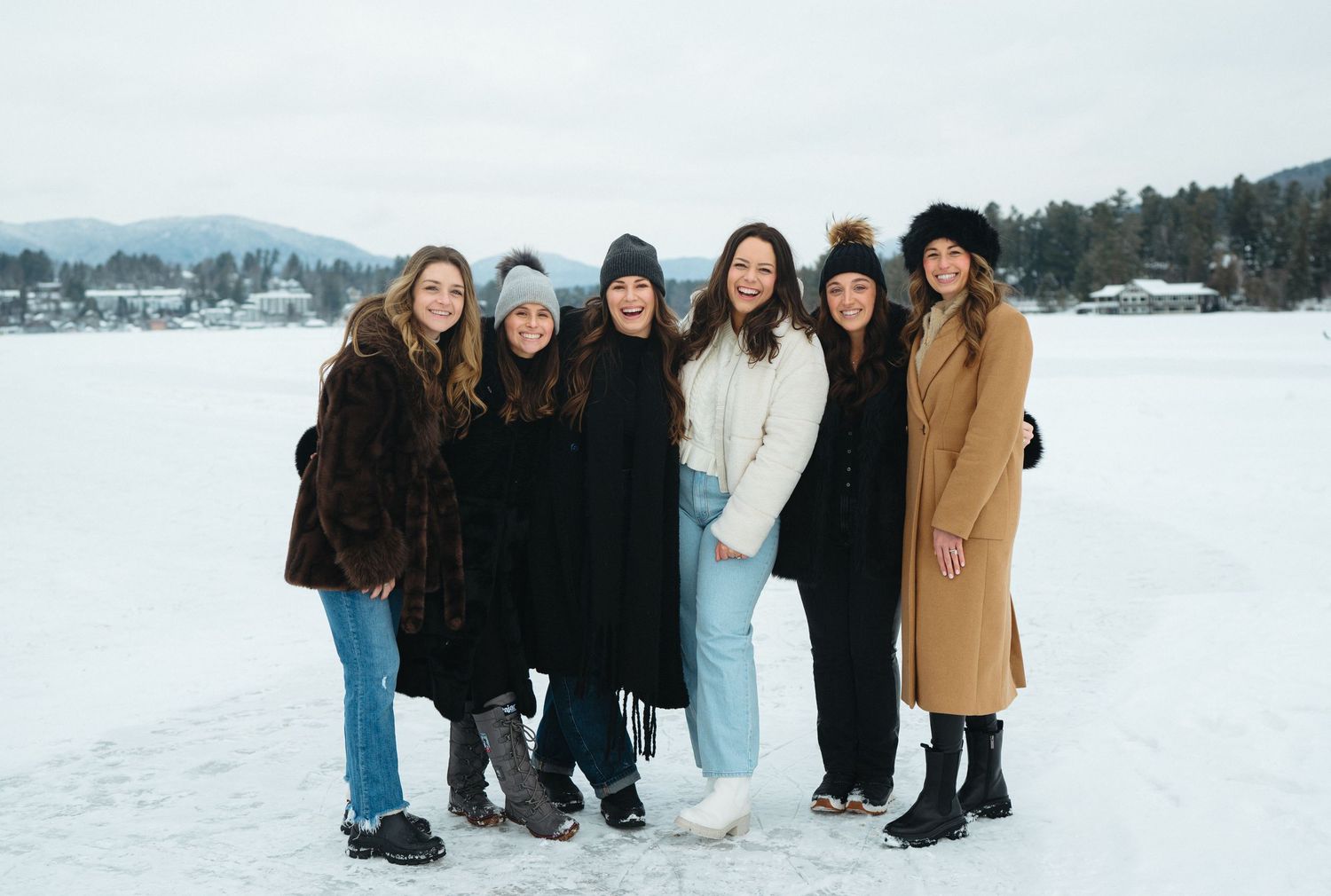 A group of friends bundled in winter coats pose together on a frozen lake with snow-covered mountains in the background.