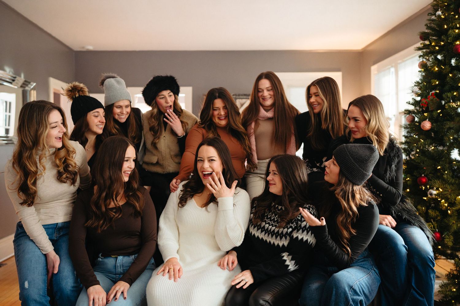 A group of friends laugh and smile together while sitting on a couch during a holiday gathering with a Christmas tree nearby.