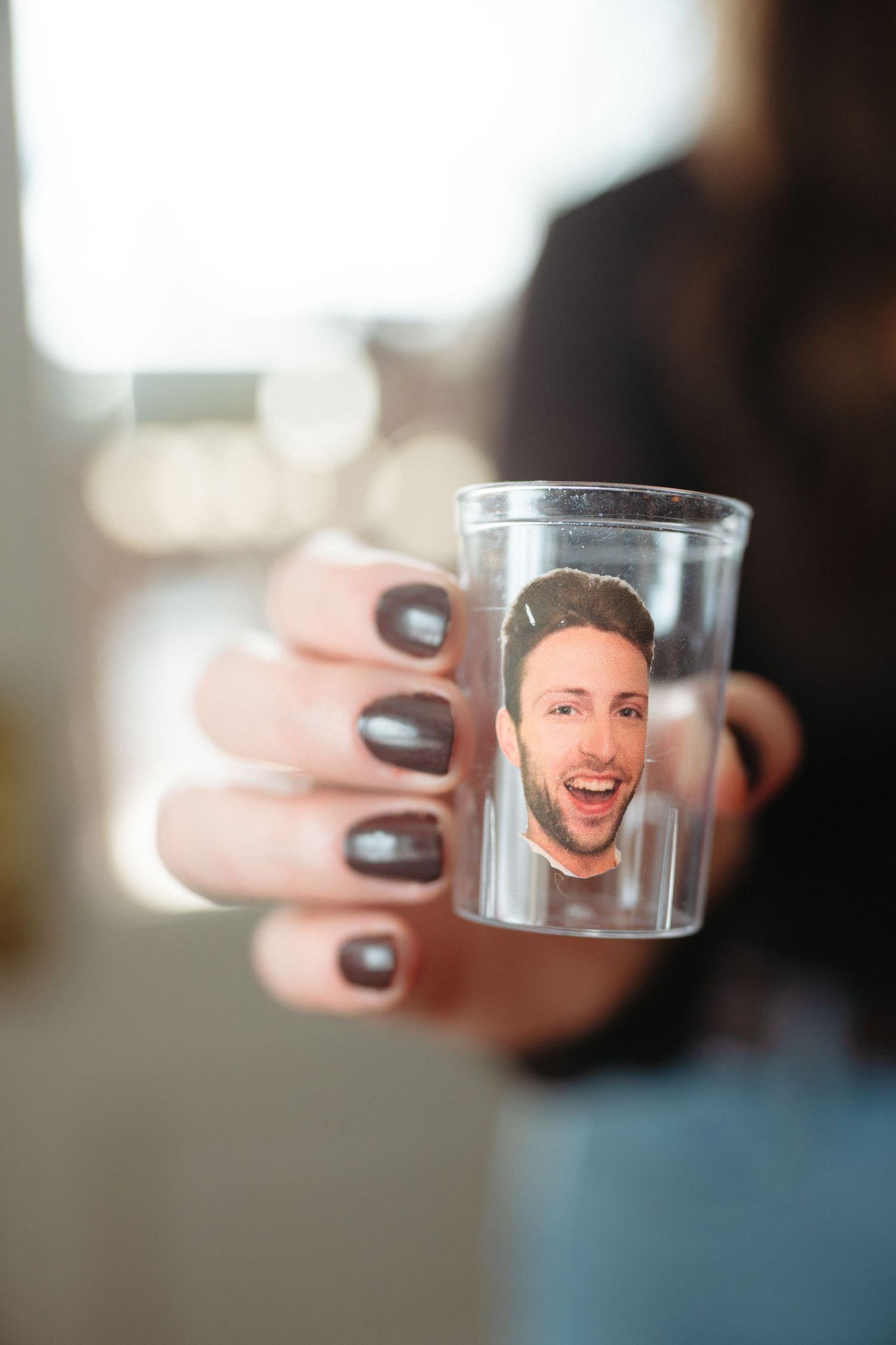 Hand with dark nail polish holding a shot glass with a photo inside against a blurred background.