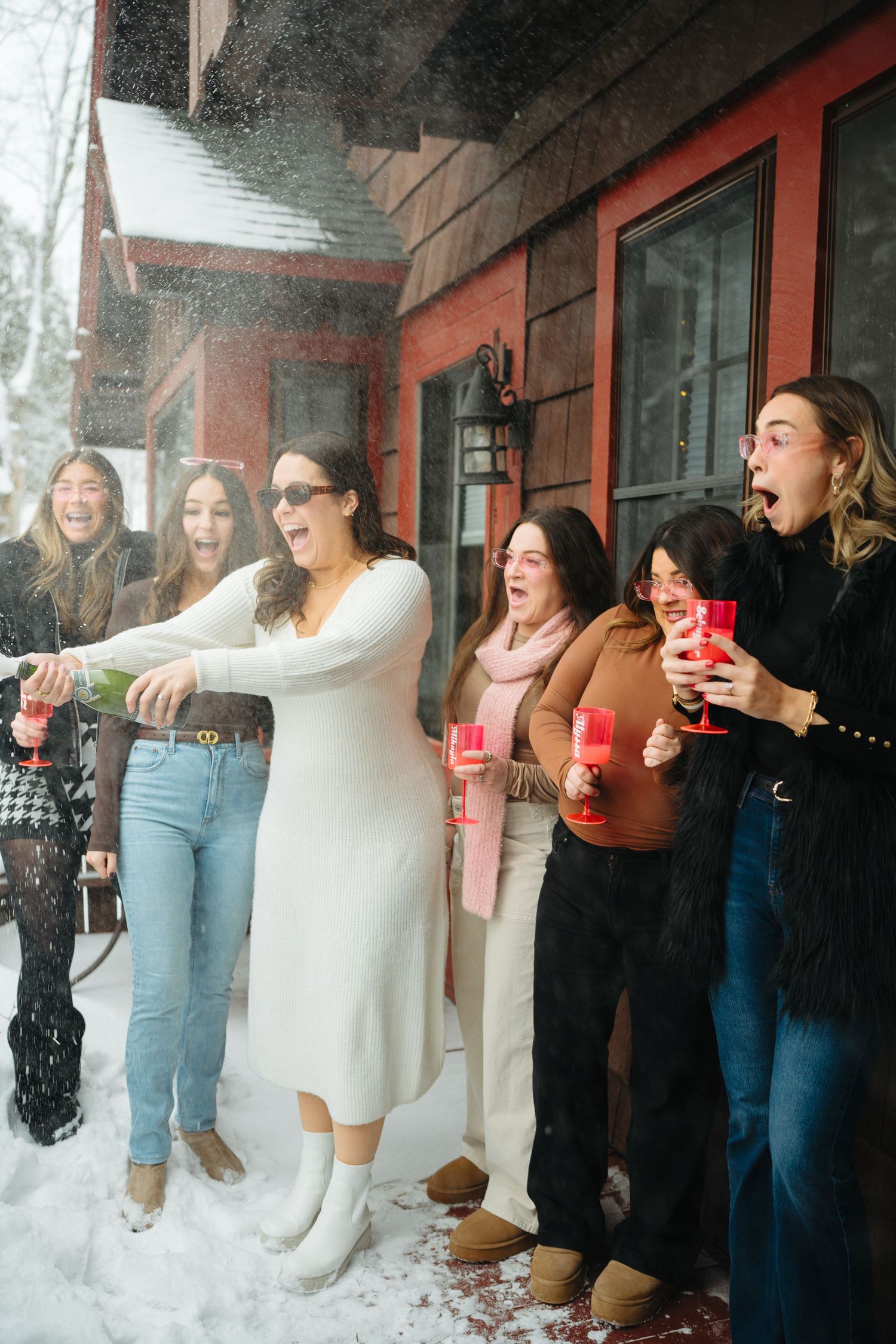 A joyful group laughs together on a snowy porch while holding red cups.