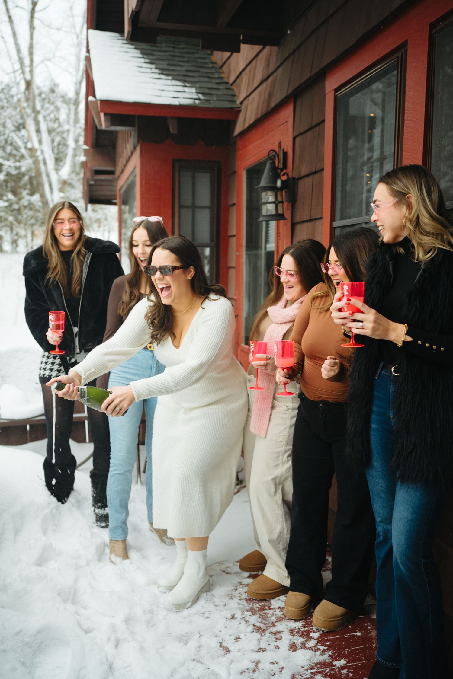 Friends gathered outside a red cabin in the snow holding beverages and laughing together.