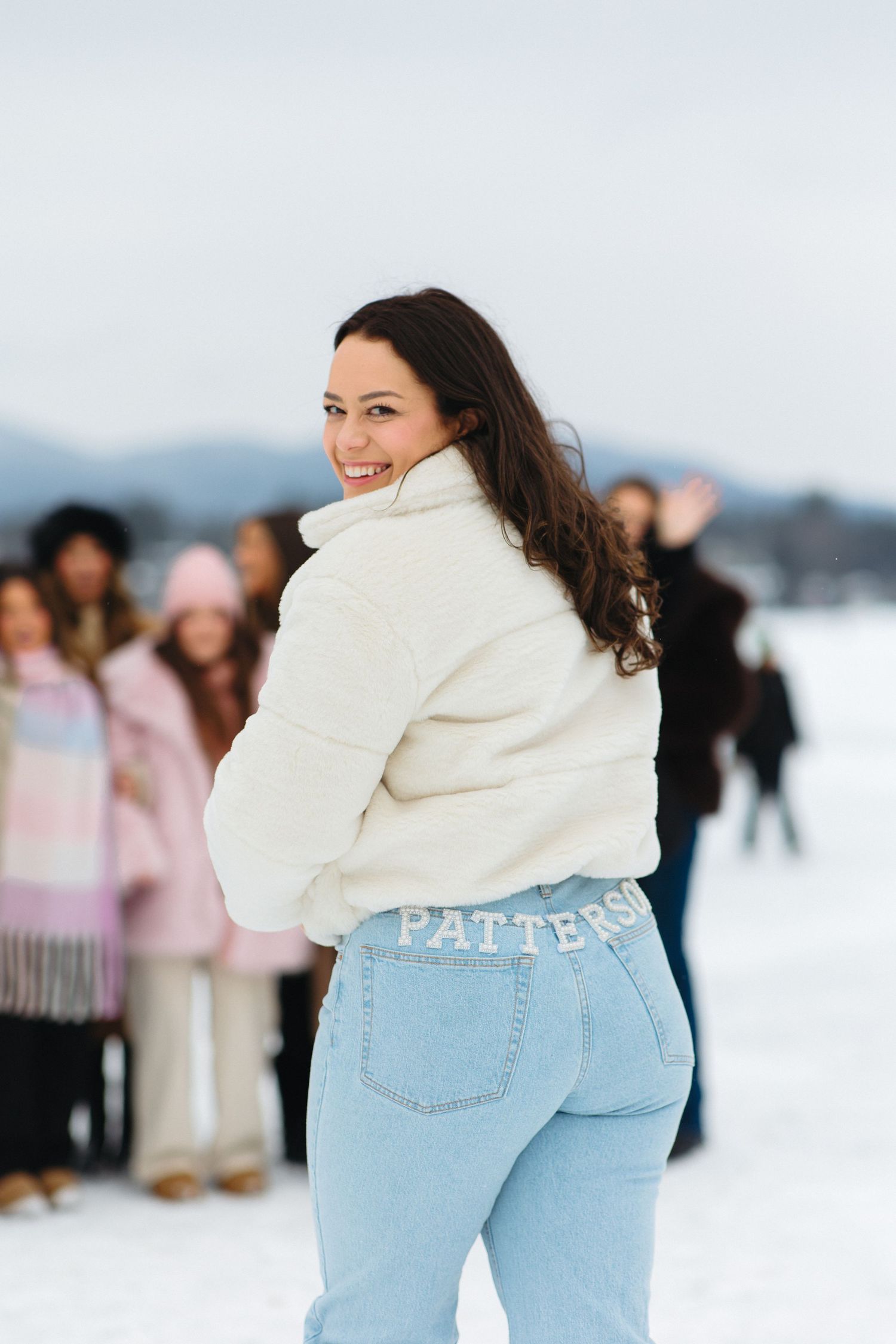 A person in a white sweater and jeans poses on a snowy winter day with others in the background.