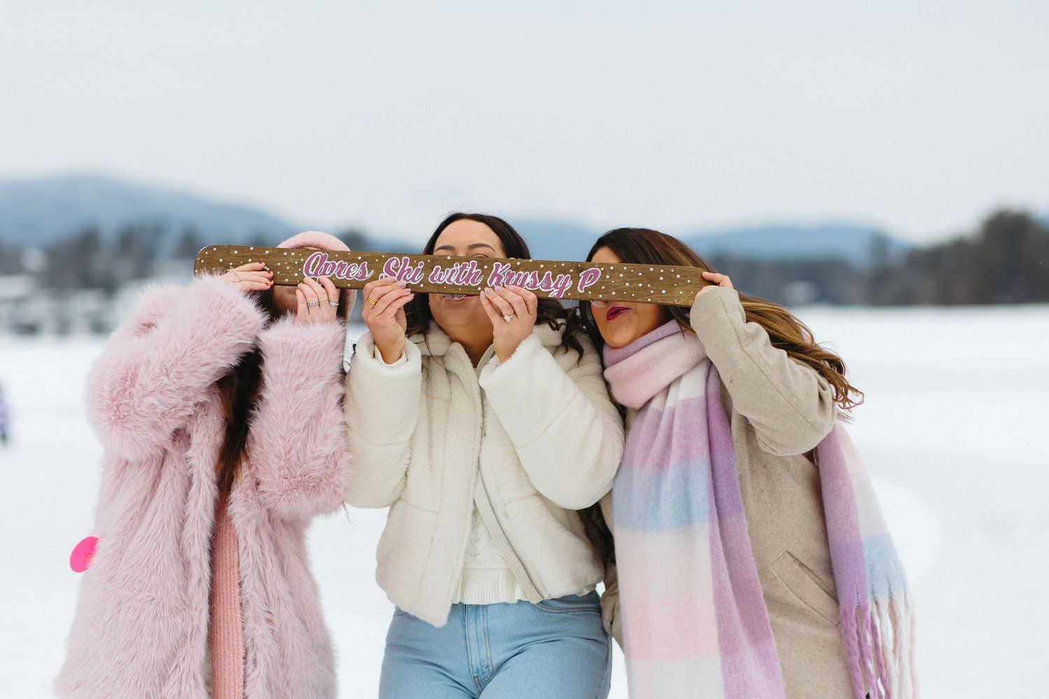 Three friends in winter coats playfully covering their eyes while standing in a snowy landscape with mountains.