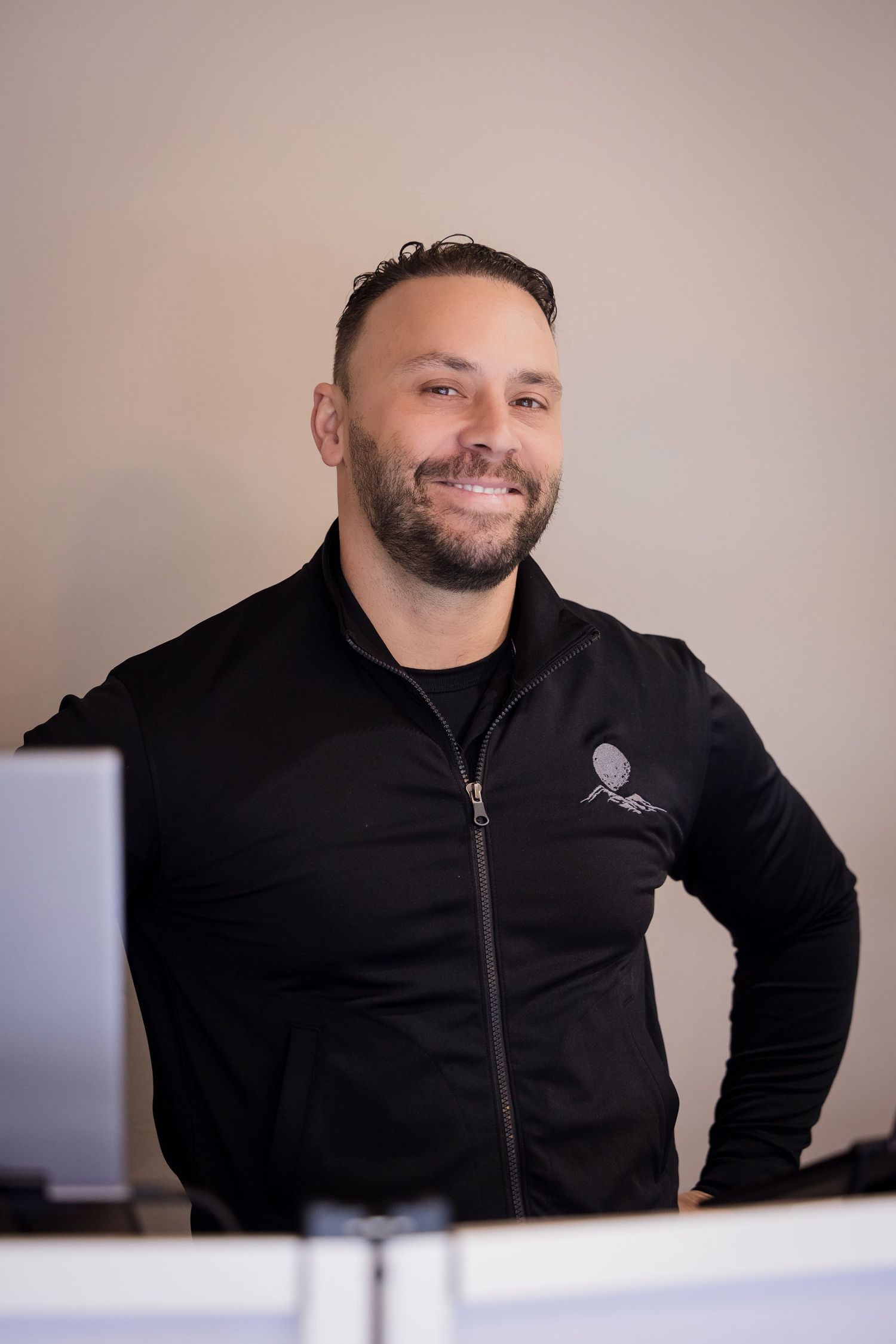 Professional in black zip-up jacket smiles warmly while sitting at desk in office setting.