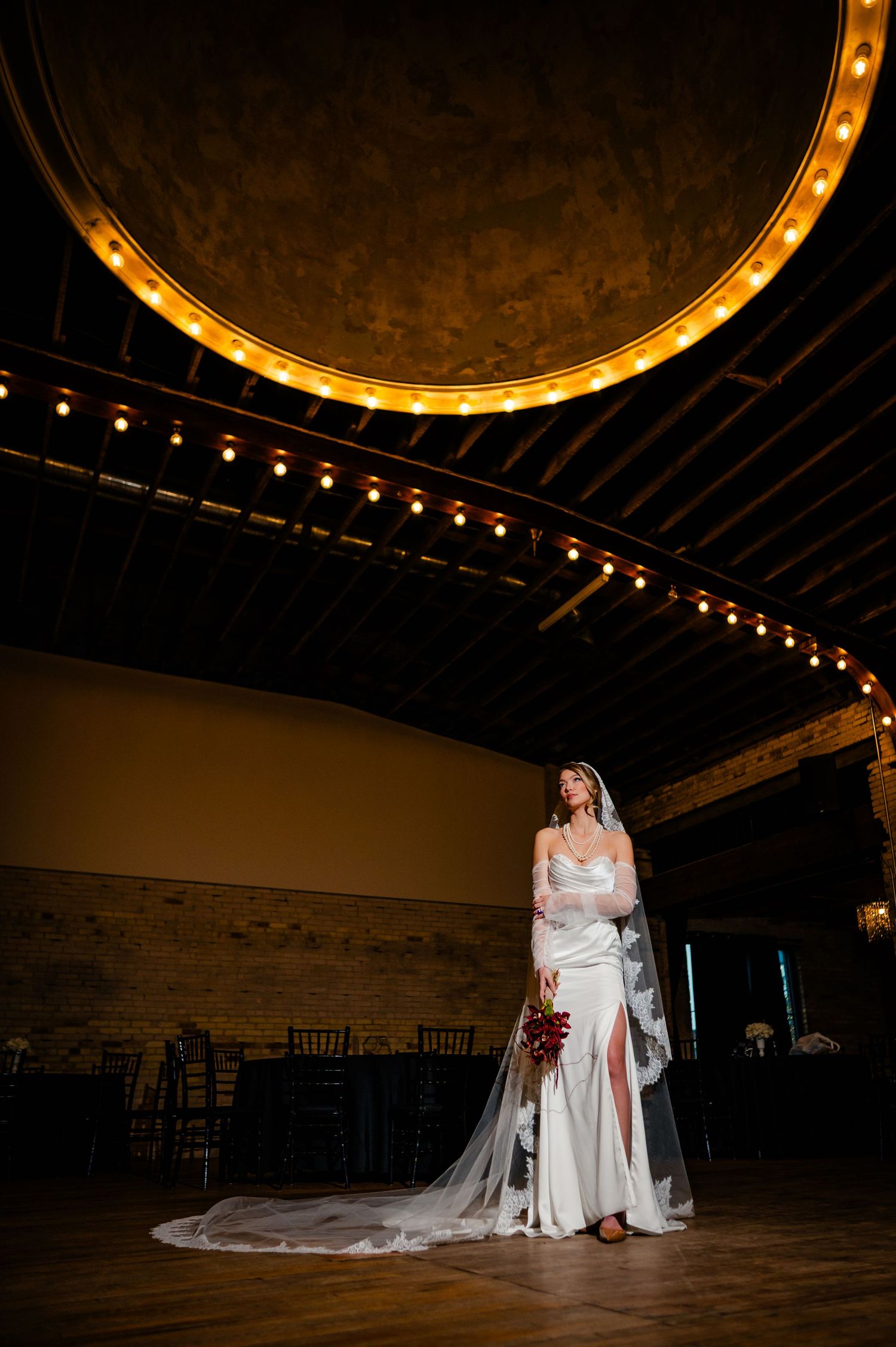 A bride in a white wedding dress sits elegantly under warm lighting in a rustic venue with string lights overhead.