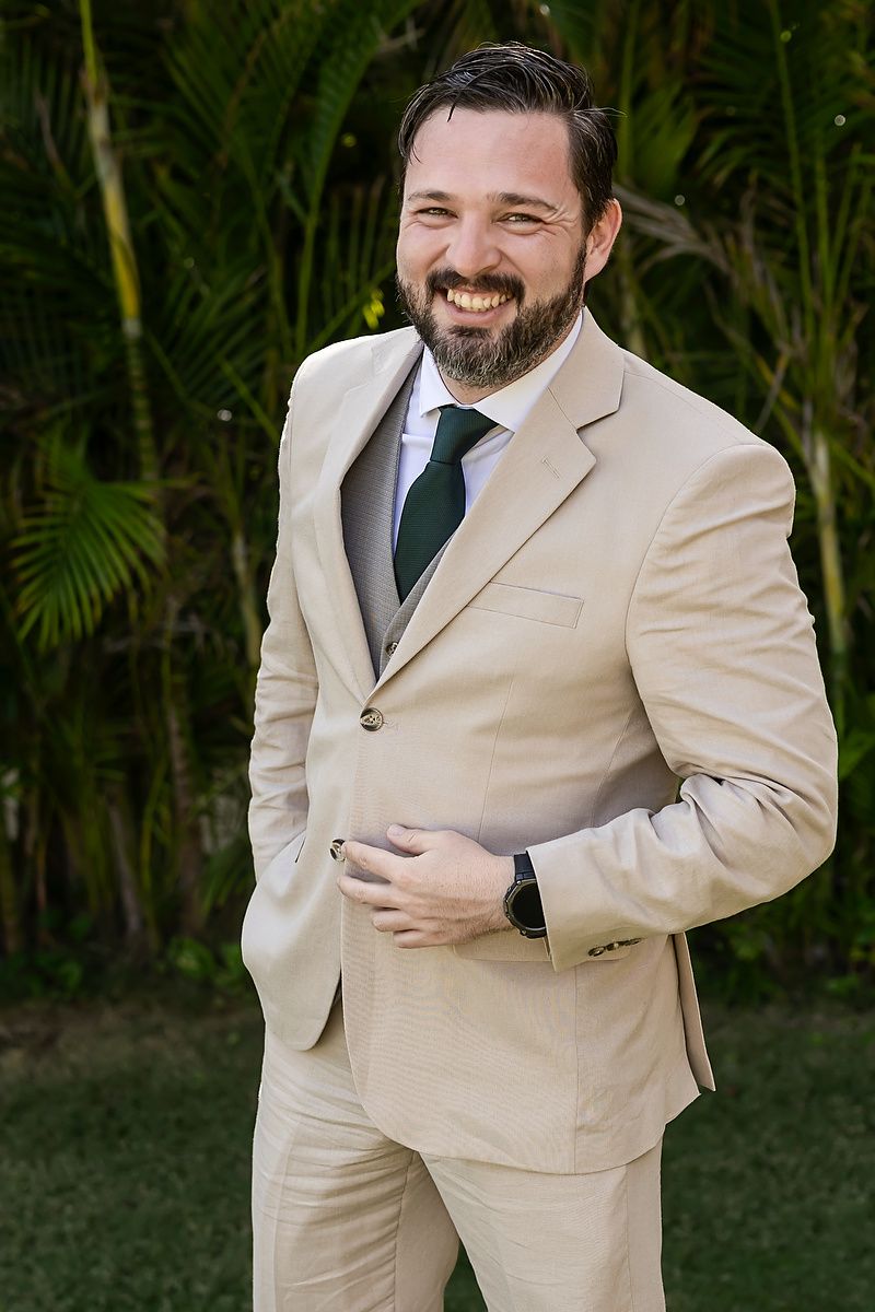 A person in a beige suit and dark tie poses outdoors with bamboo plants in the background.