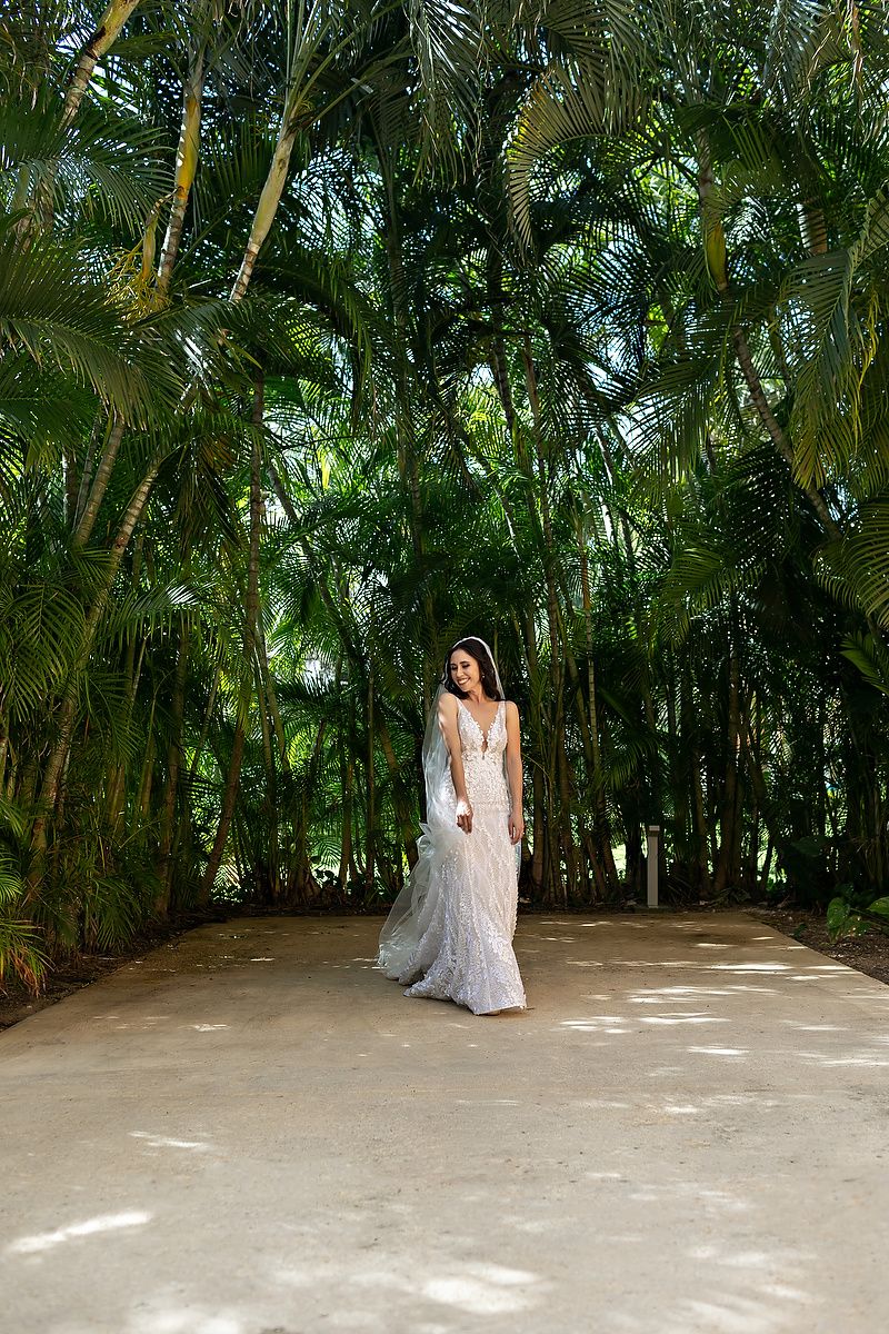 A bride in a white wedding dress poses in a lush tropical garden setting with tall palm trees surrounding her.