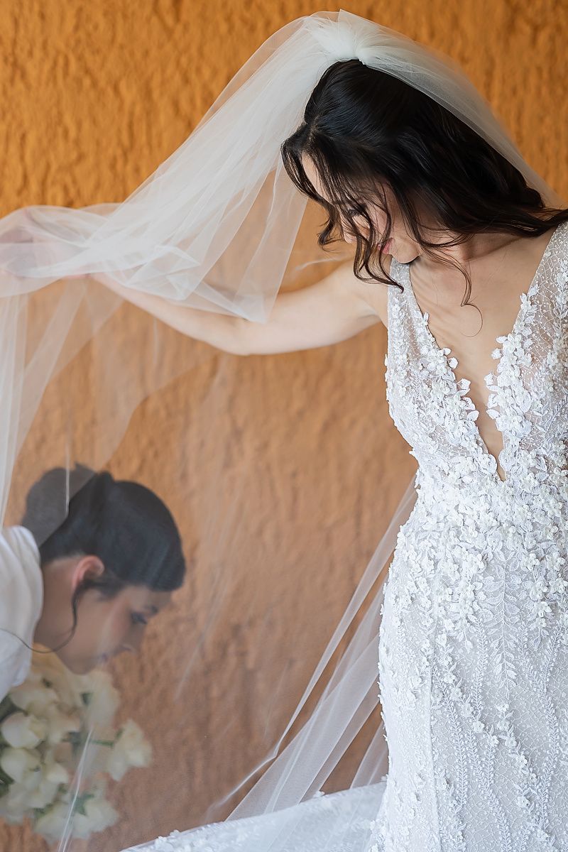 A bride in a white lace wedding dress with a flowing veil stands against a textured orange wall.