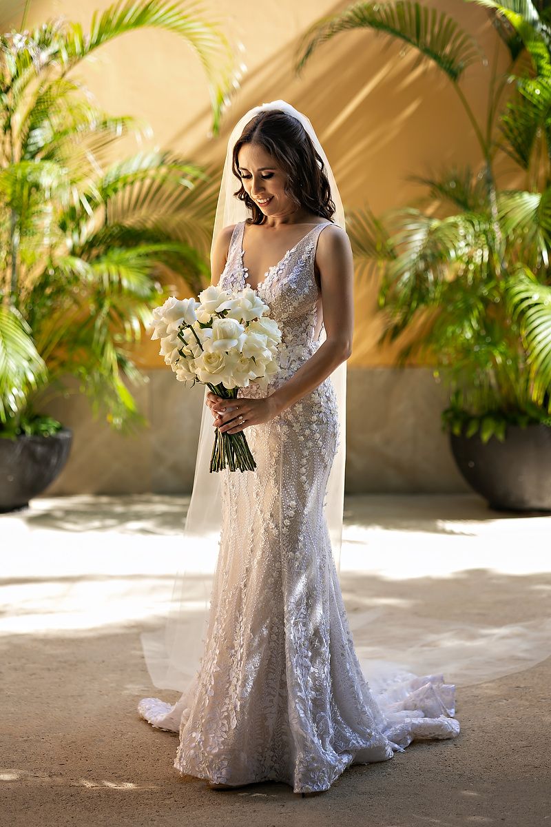 Elegant white lace wedding dress with flowing train and bouquet captured in sunlit tropical garden setting.