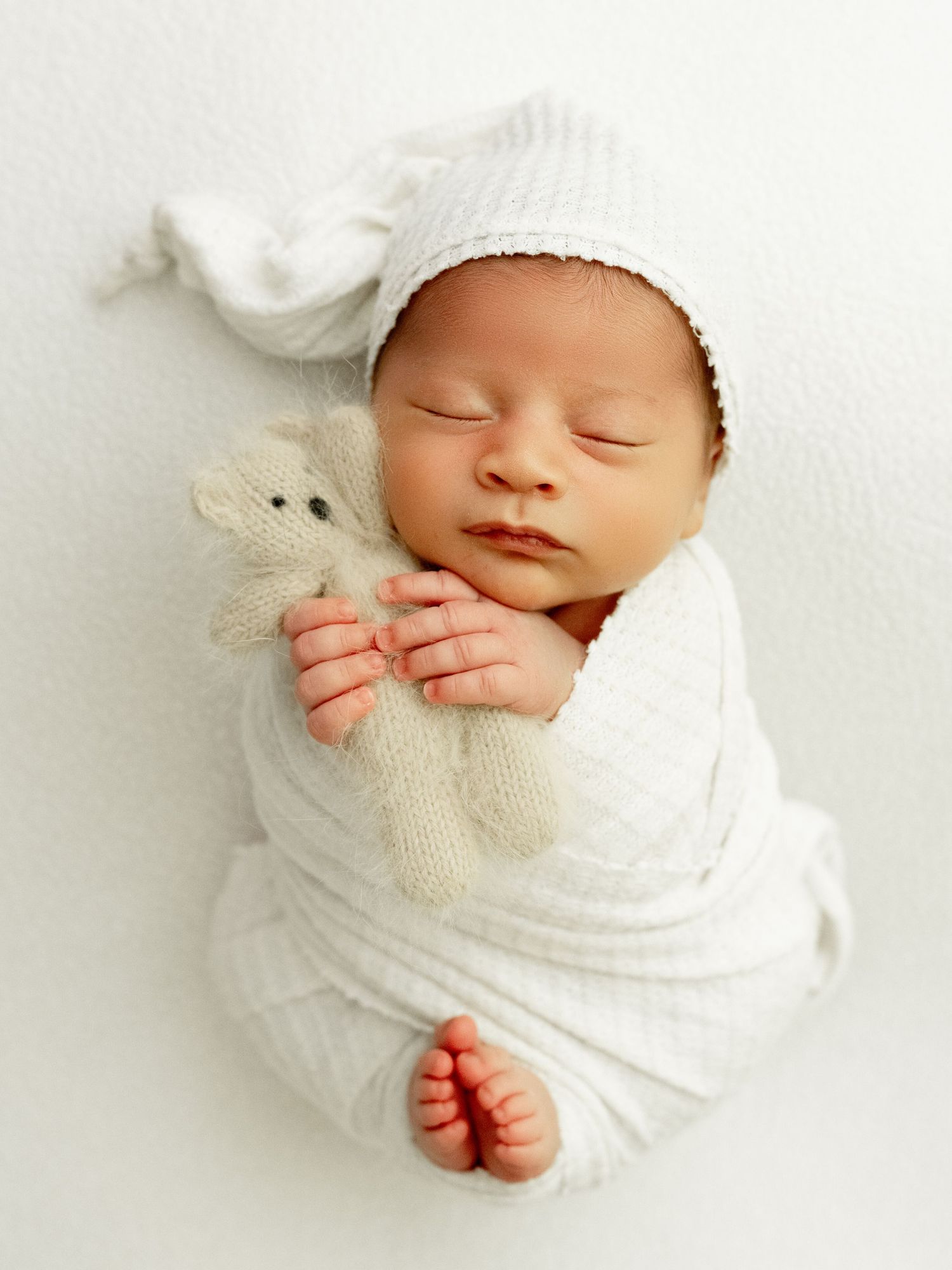 Parent in gray sweatshirt tenderly holds and kisses sleeping newborn baby in cozy neutral studio setting.