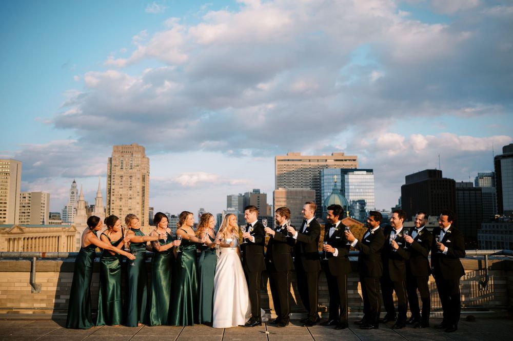 Wedding party lined up on rooftop with city skyline backdrop at sunset.