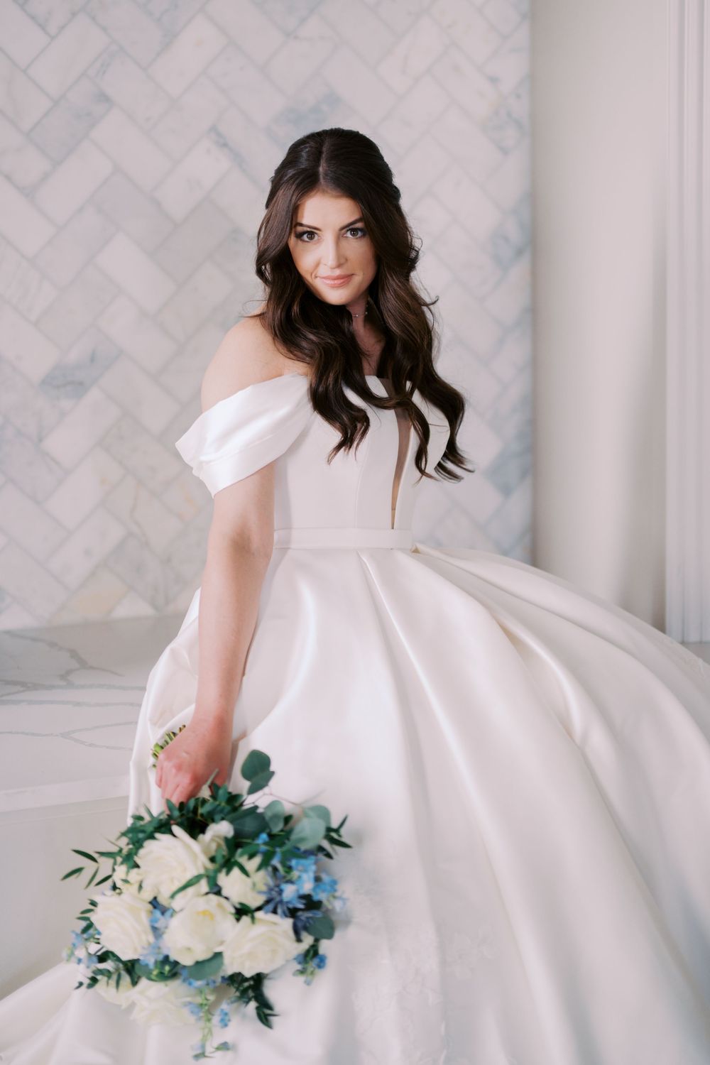 A bride in a flowing white dress holds a blue and white floral bouquet against a light gray patterned wall.
