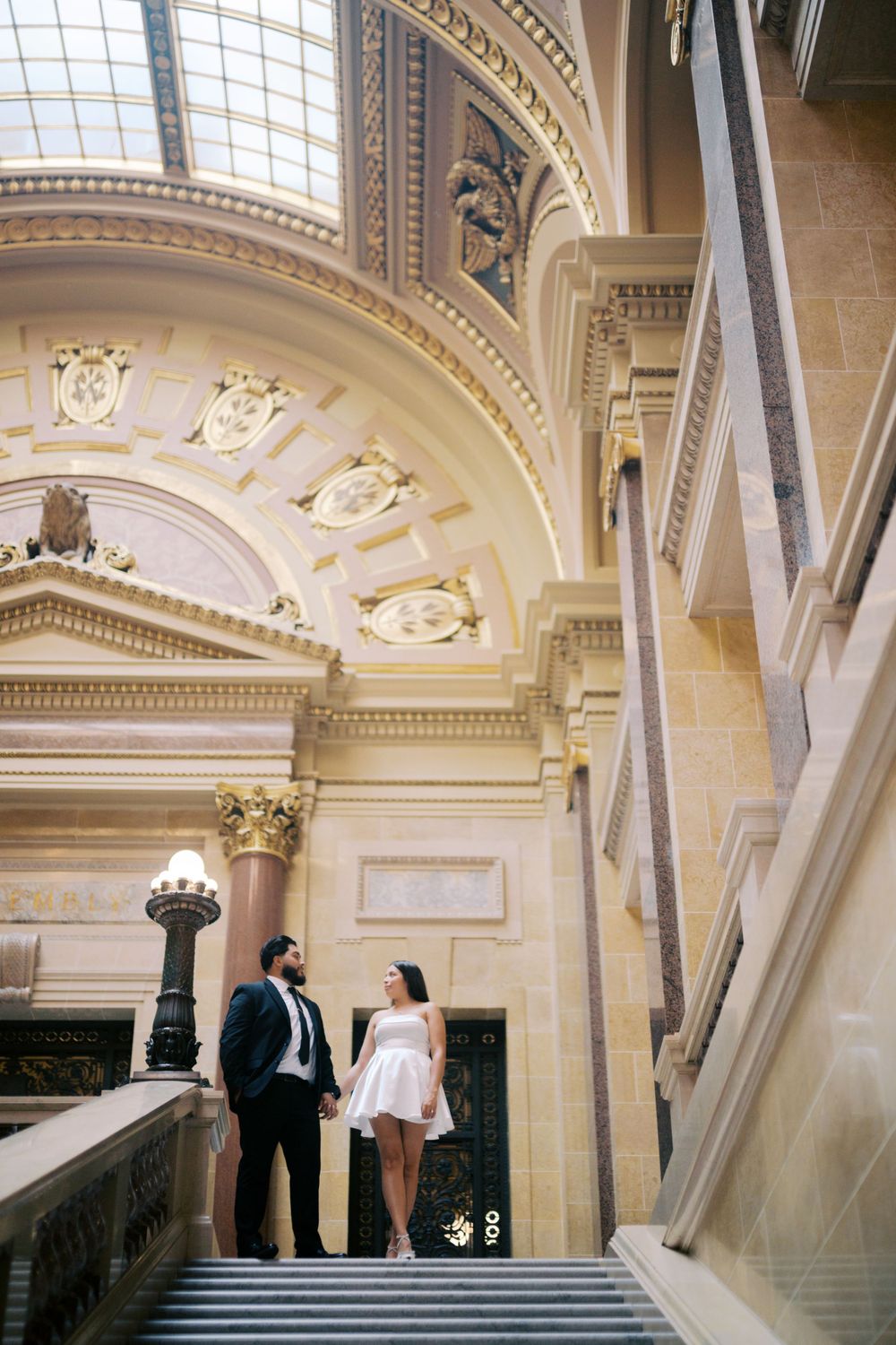 Couple poses on grand staircase beneath ornate architectural dome and columns.