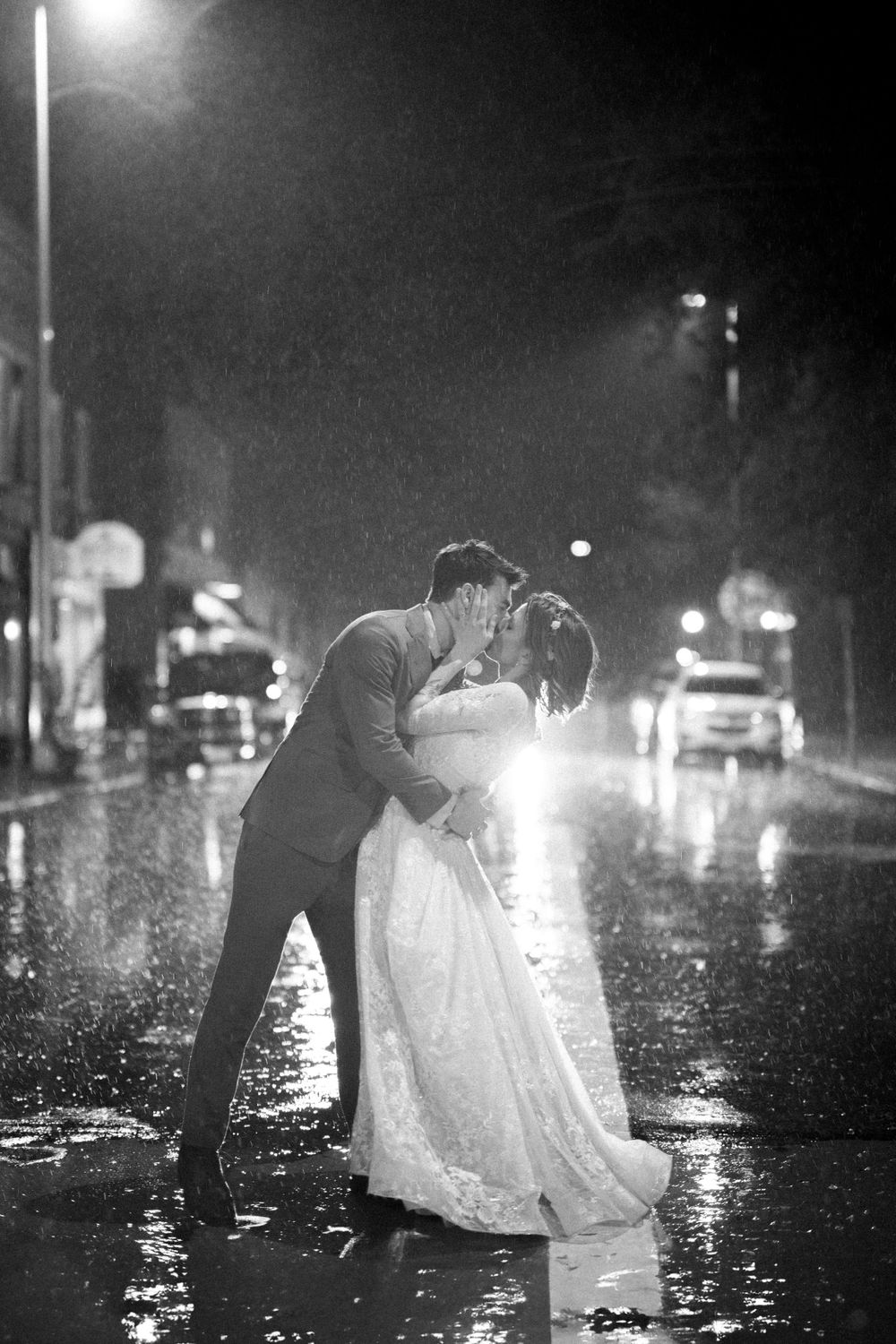Romantic rainy night wedding portrait with couple kissing under streetlights.