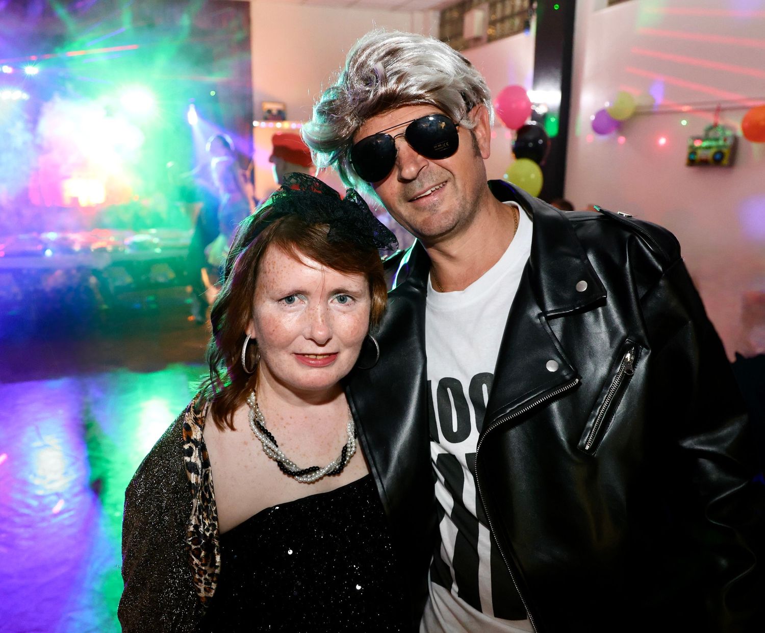 A couple in festive attire poses together at a party with colorful disco lights in the background.
