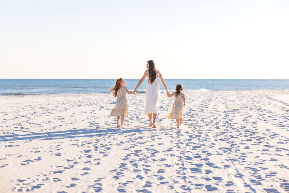 Three figures in white dresses walk hand in hand on a pristine sandy beach with ocean waves in the background.