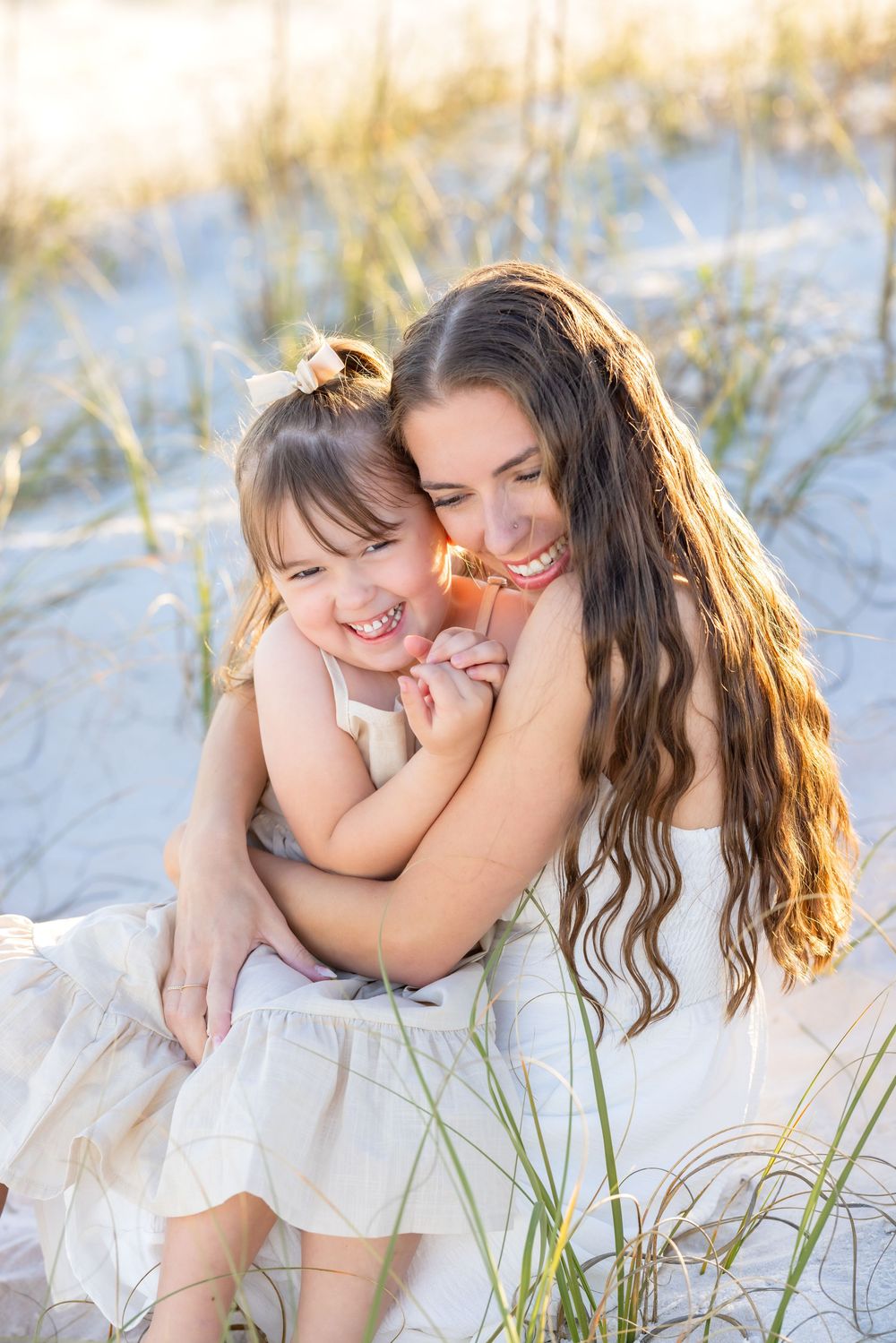 A tender moment captured in white outfits against beach sand dunes and sea grass at sunset.