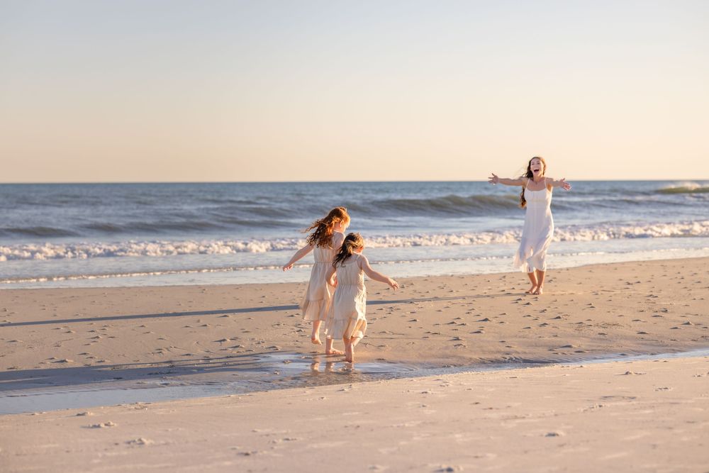 Two people in white dresses run joyfully along a sandy beach at sunset with ocean waves in the background.