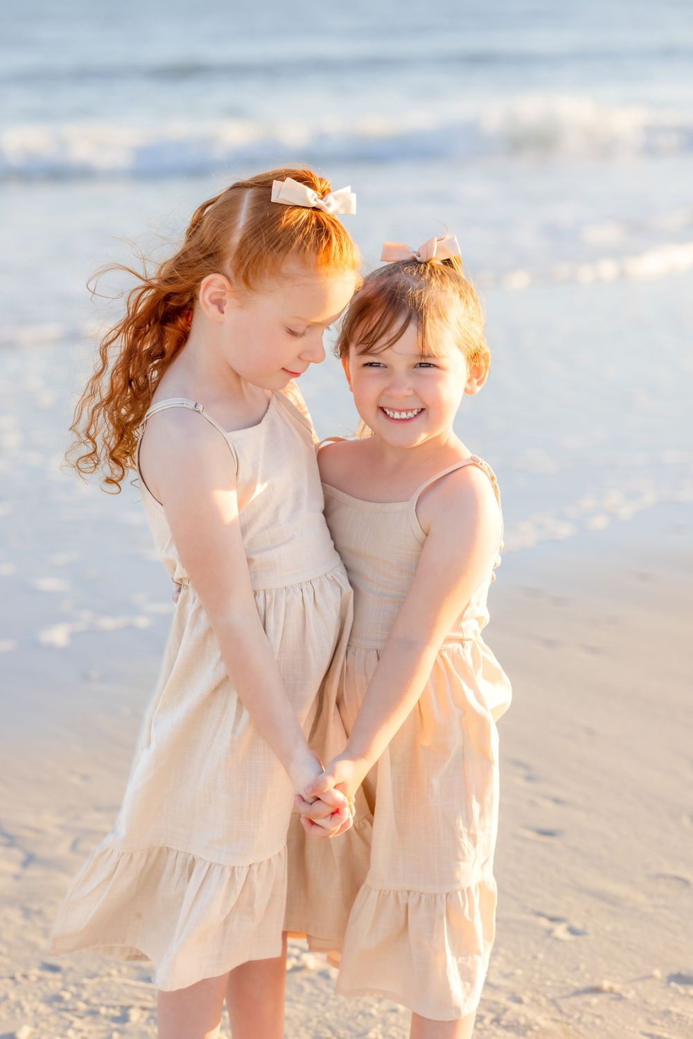 Two young children in matching beige dresses hold hands and laugh together on a sunny beach.
