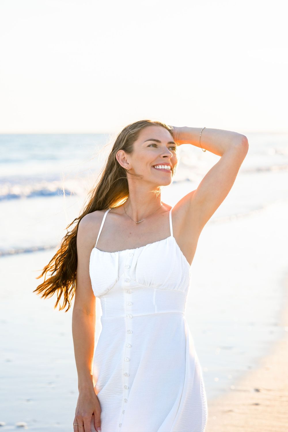 A woman in a flowing white dress walks along a sunny beach, her hair blowing in the ocean breeze.