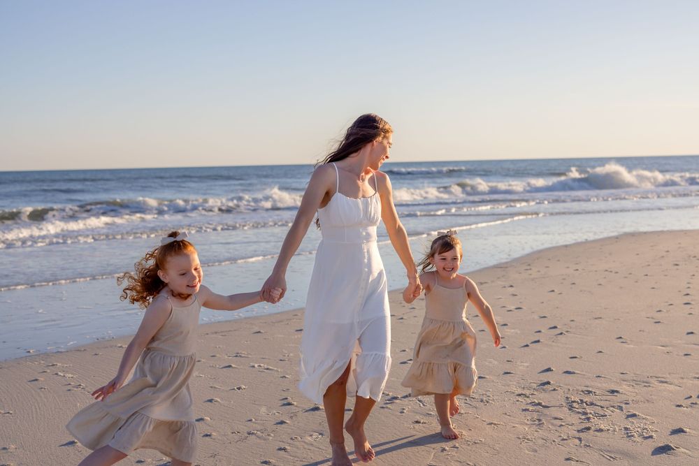 Three people in white dresses run and play together along a sunny beach shoreline at sunset.