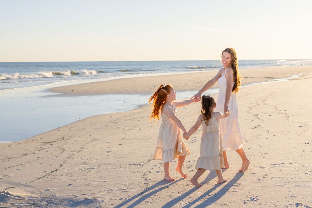 Three figures in light dresses walk hand in hand along a serene beach at sunset casting long shadows on the sand.