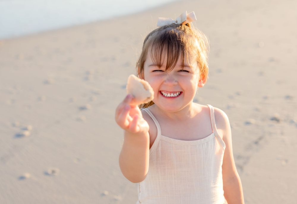 A child in a white dress smiles and poses joyfully on a sunny beach.