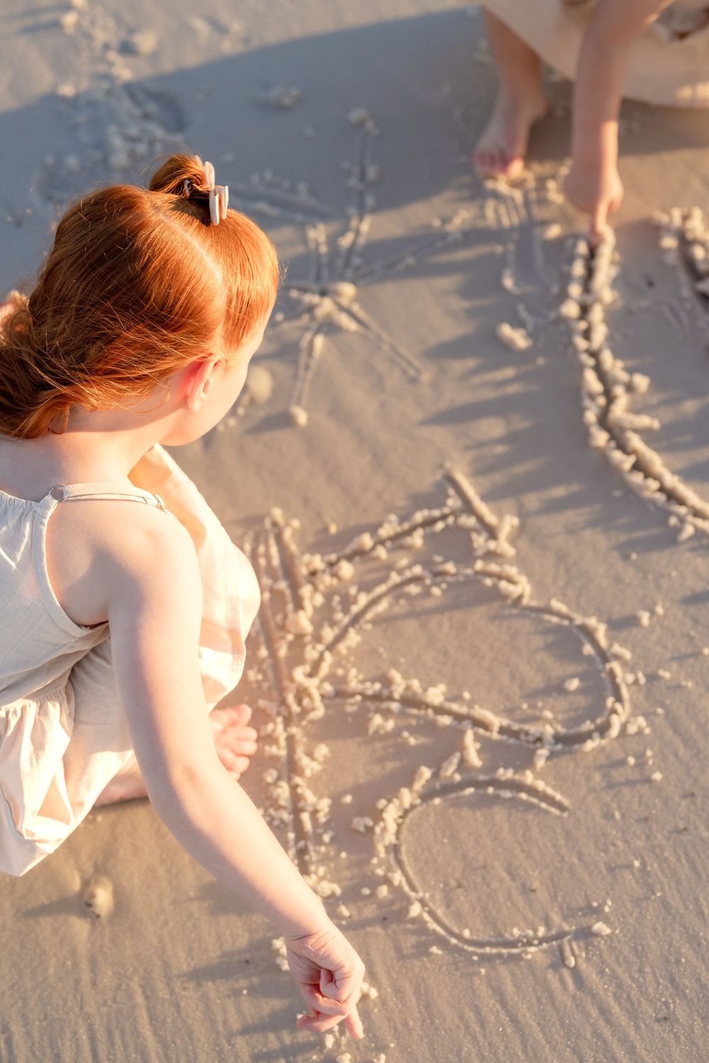 A person in white clothing draws heart shapes in the sand on a sunny beach.