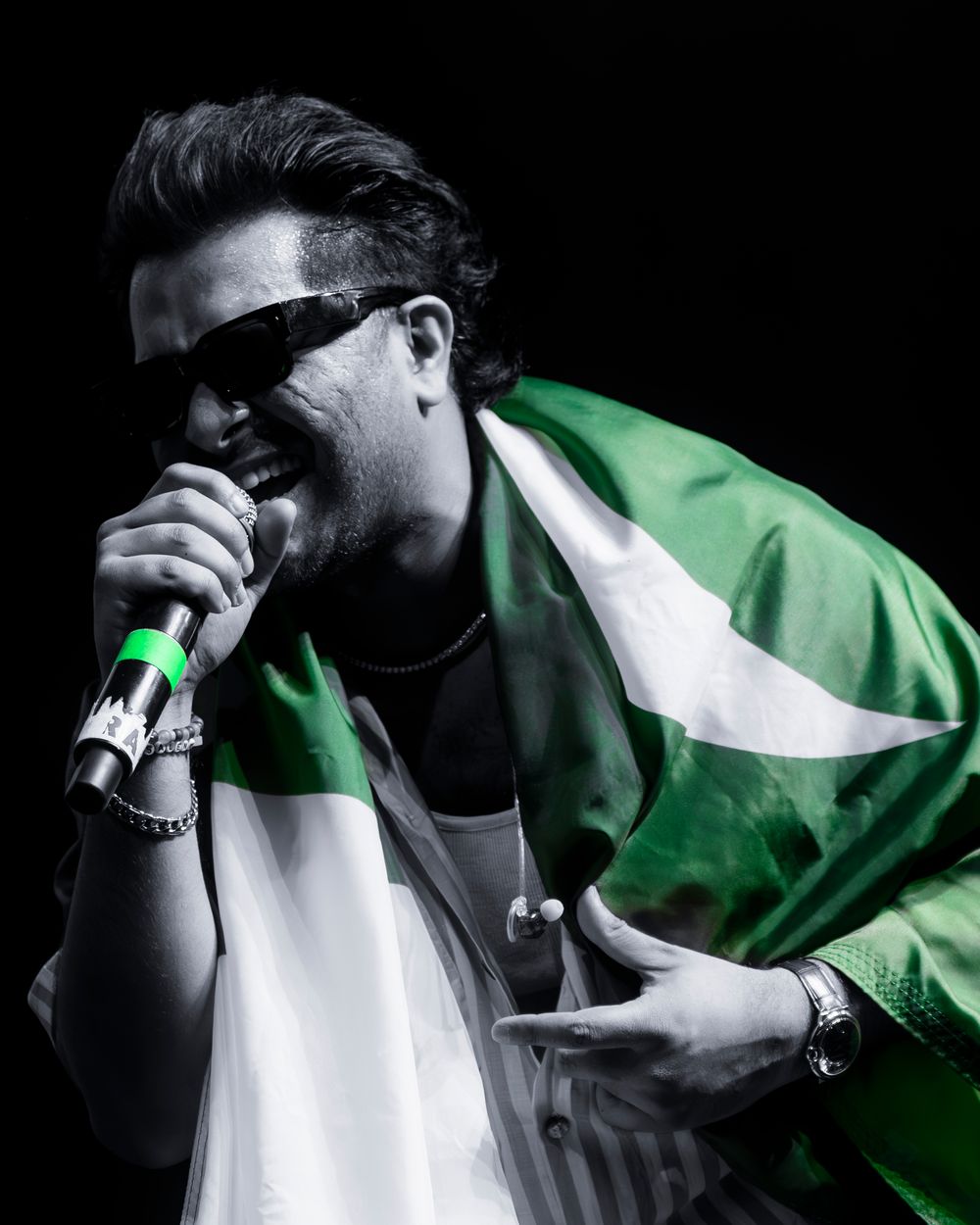 Man singing into microphone draped in Pakistani flag, wearing sunglasses, performing live on stage in black and white photo.