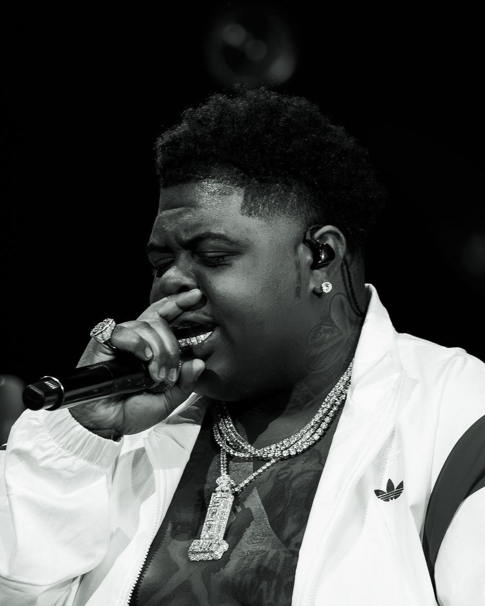 Black and white photo of a young male rapper performing on stage, singing into a microphone wearing chains and Adidas jacket.