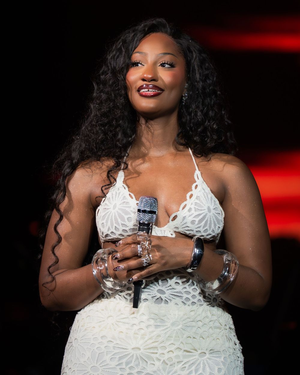 Woman in white lace dress holding microphone on stage, wearing bracelets and rings, with long curly hair.