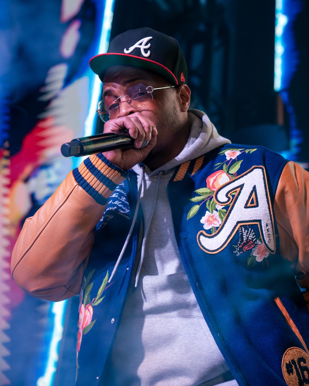Performer wearing Atlanta Braves baseball cap and floral varsity jacket holds microphone on stage with blue lighting.