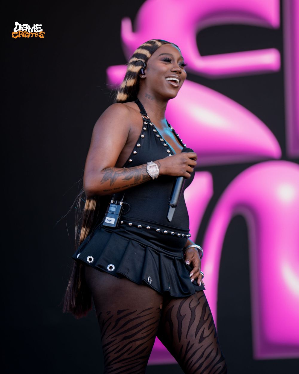Performer in black dress and brown pants dances energetically on stage with vibrant pink lighting in the background.