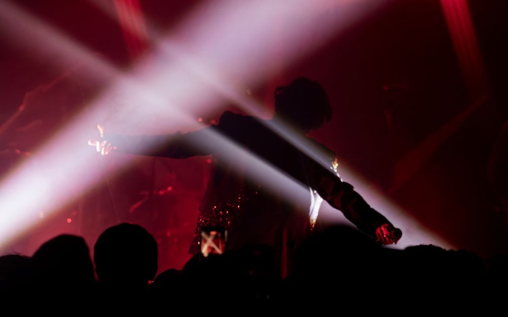 Concert crowd silhouetted against dramatic red and white stage light beams at a live music event.