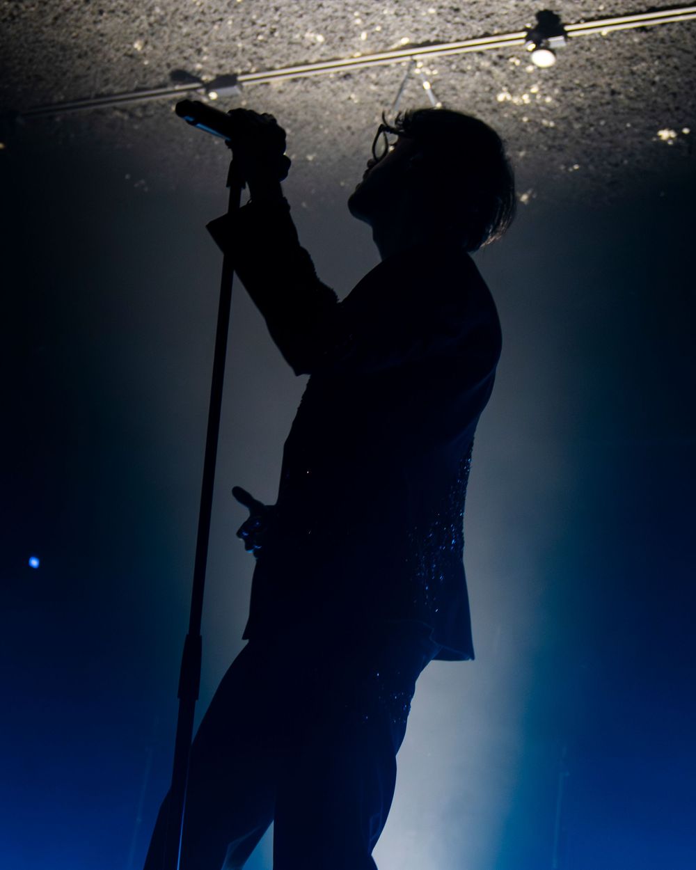Silhouette of a male singer performing on stage, holding a microphone stand with dramatic blue backlit lighting.