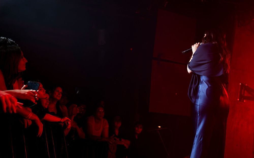 Performer on stage with microphone in dimly lit red venue with audience watching live music show.