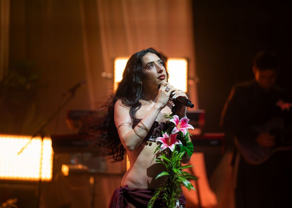 Woman singer holding pink lilies on stage, performing under warm dramatic lighting at a live concert.