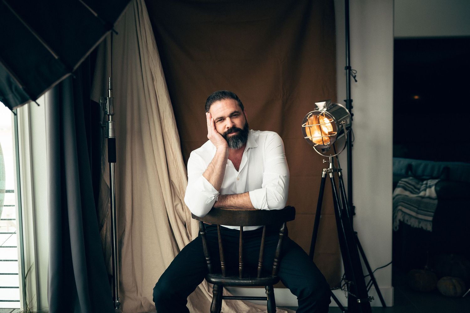 Bearded man in white shirt sitting on chair with hand on cheek in a warmly lit studio setting.