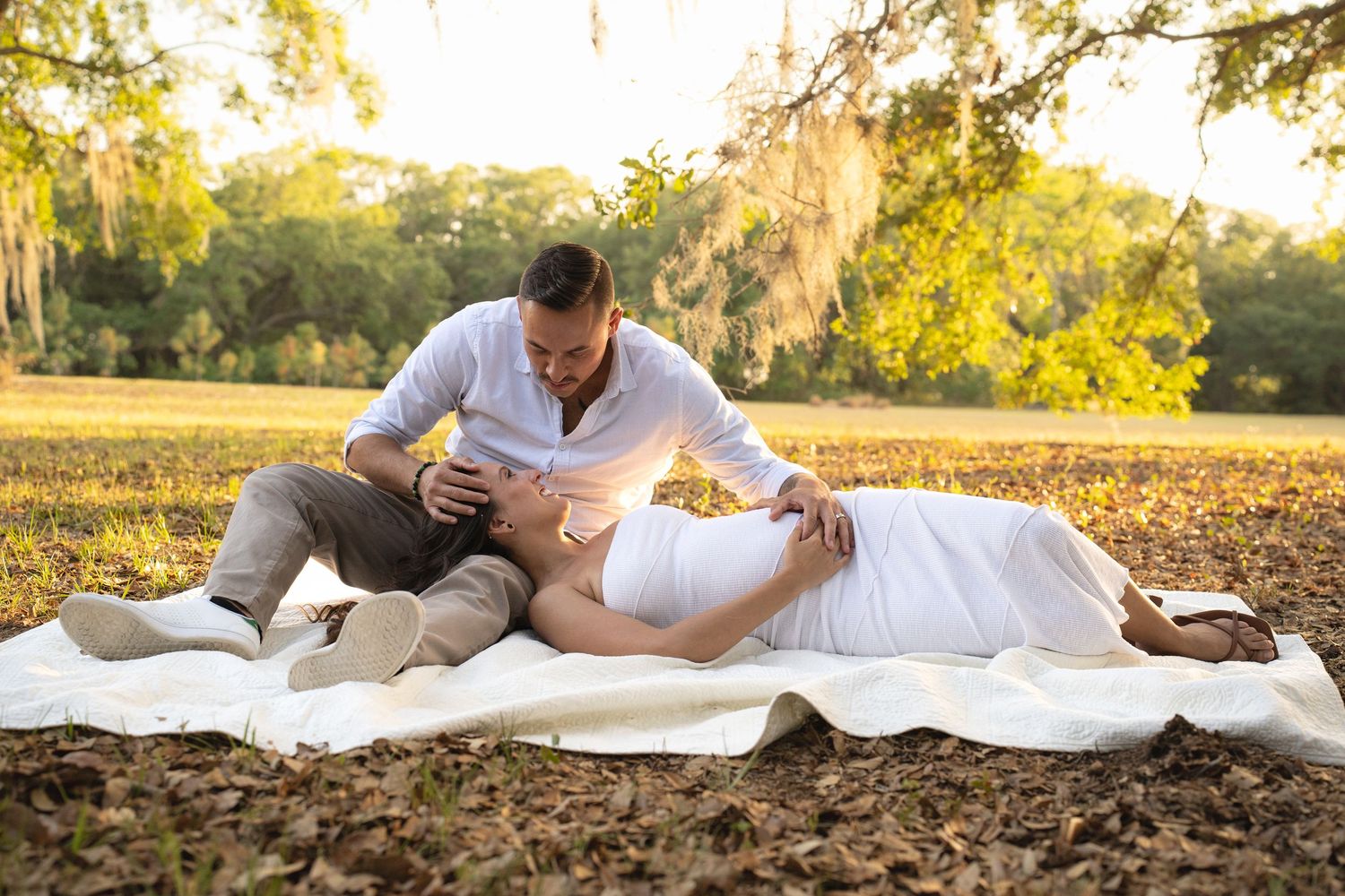 Engaged couple capture this lovely moment at sunset on a picnic blanket in a warm park.