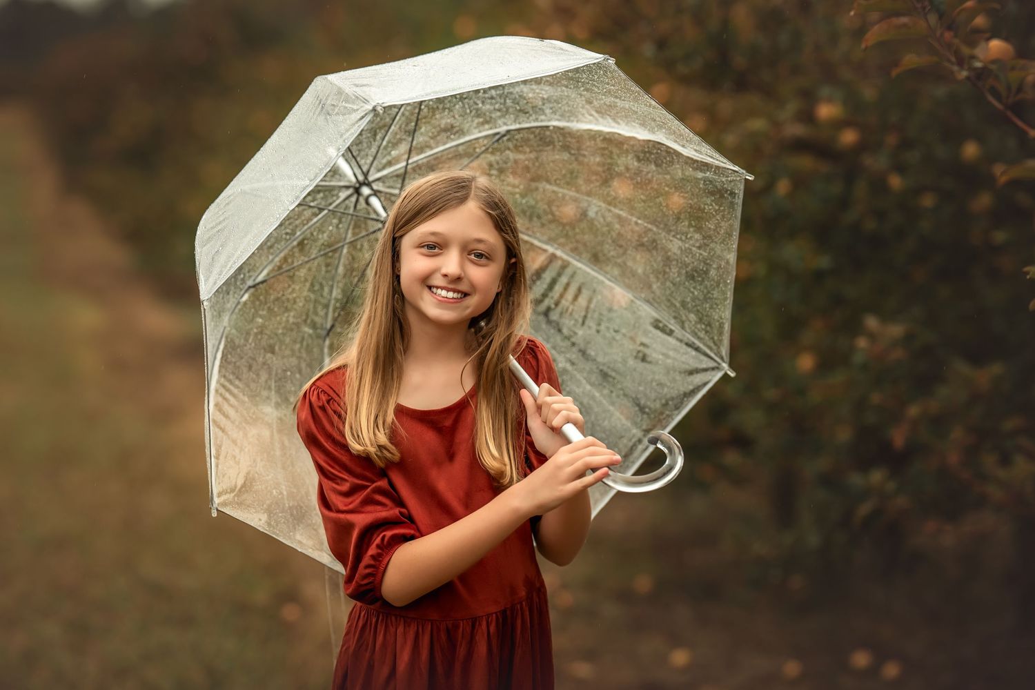 A person in a burgundy dress holds a clear umbrella while standing outdoors in autumn weather.