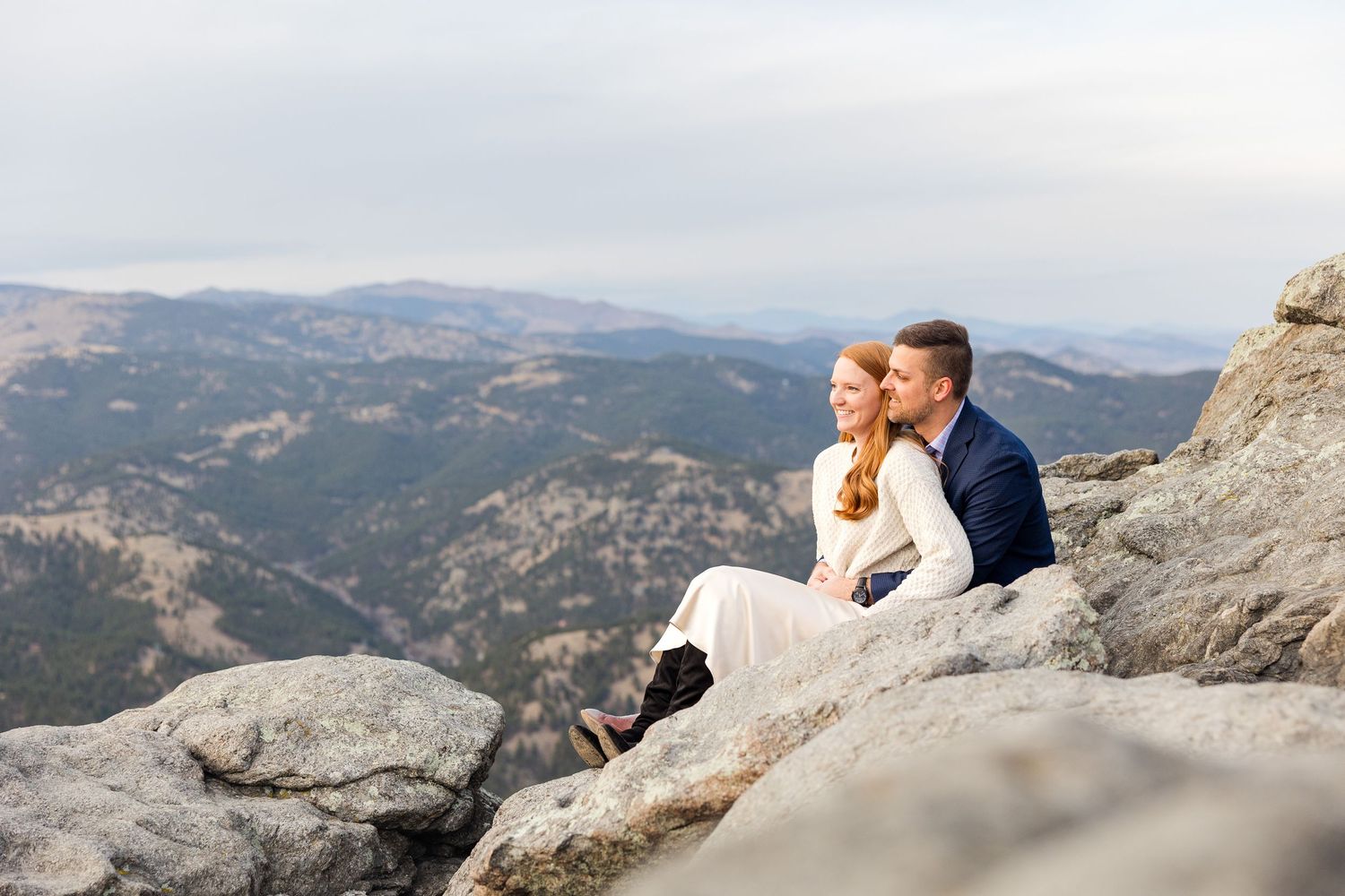 A couple in wedding attire sits together on a mountain peak overlooking a scenic valley and distant mountain ranges.