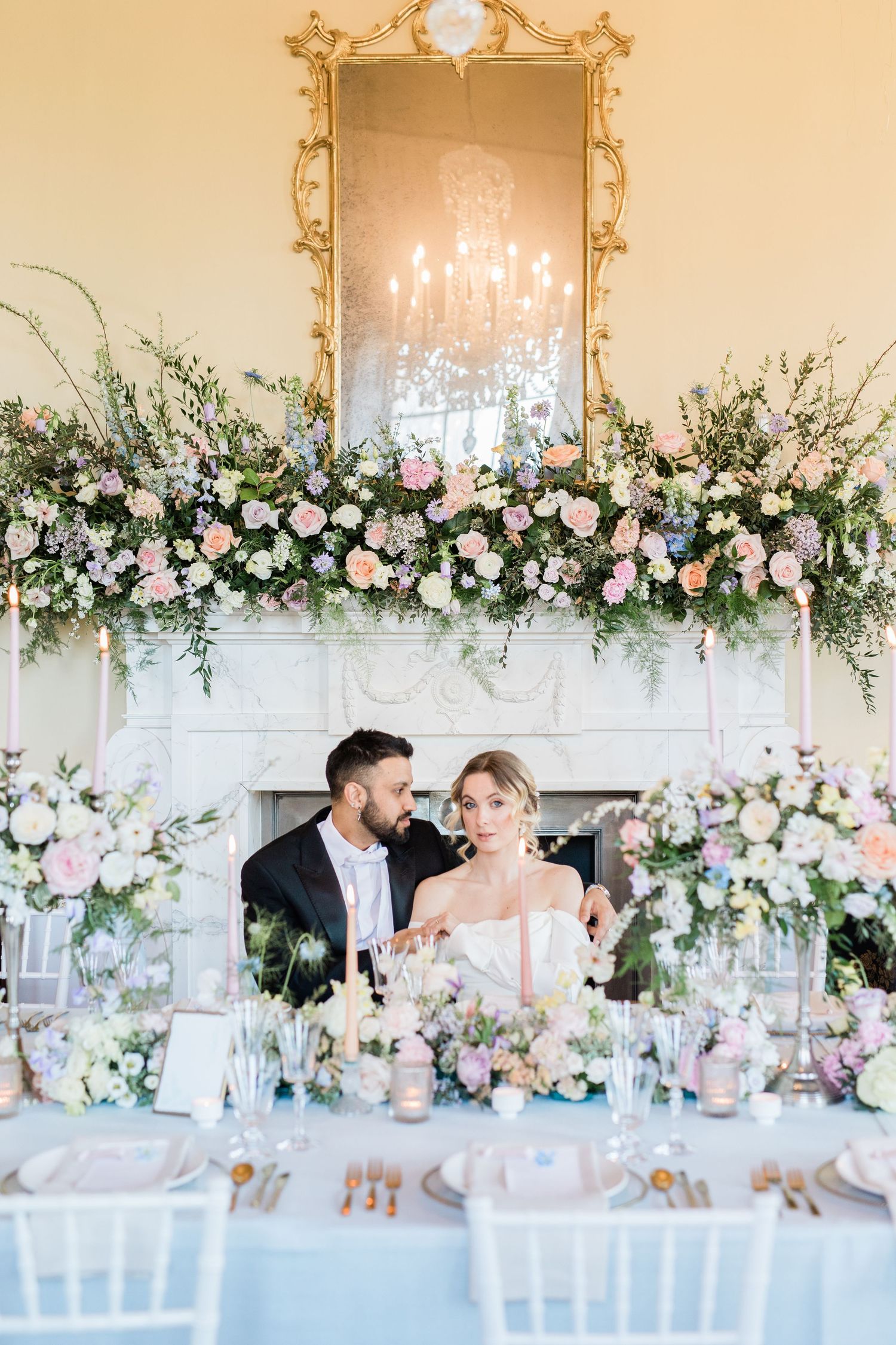 Elegant wedding reception table with white floral garlands, candelabras and pastel decor against a light neutral wall.