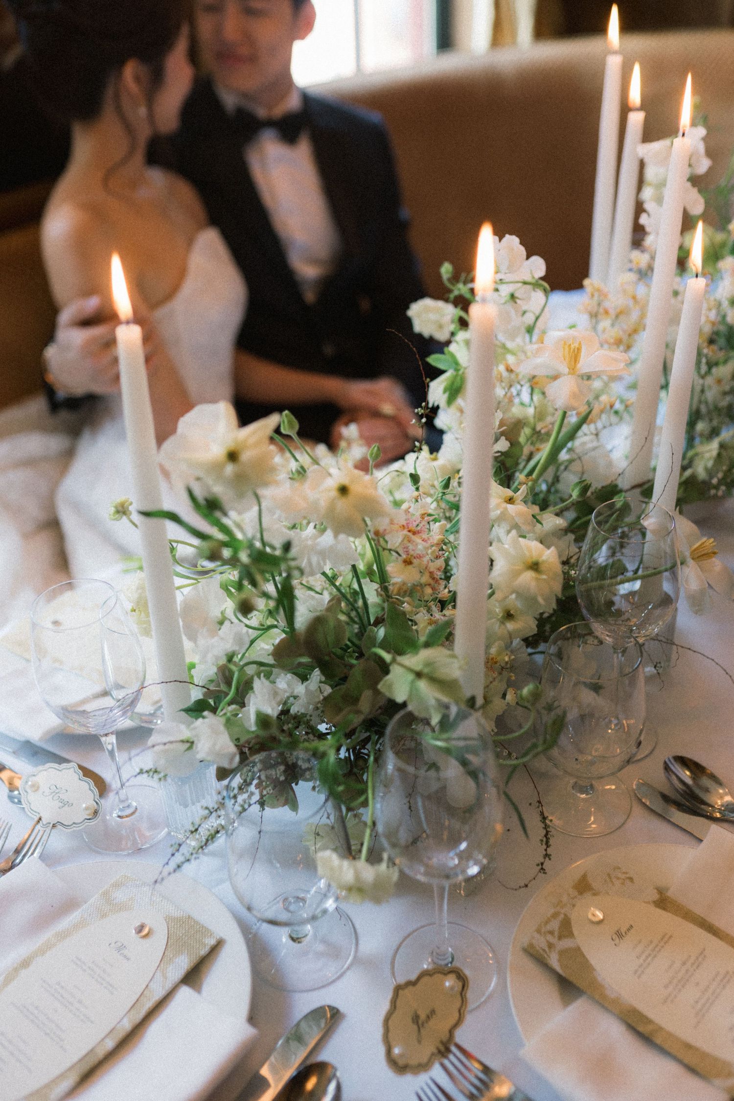 Elegant wedding table with white floral centerpiece, tall taper candles, crystal glasses, and couple in background.