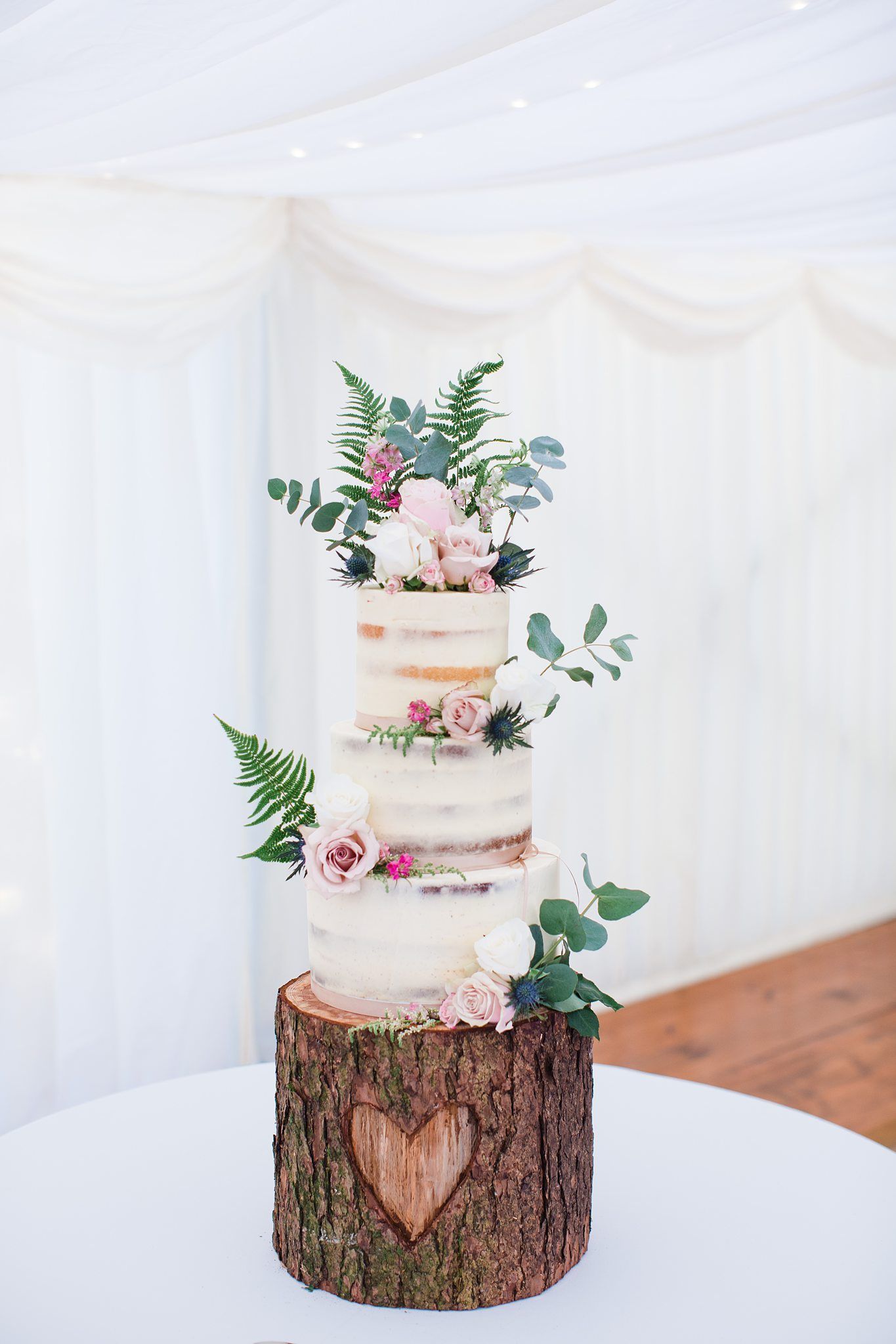 Three-tier semi-naked wedding cake with pink roses and ferns on a rustic wood log stand with carved heart.