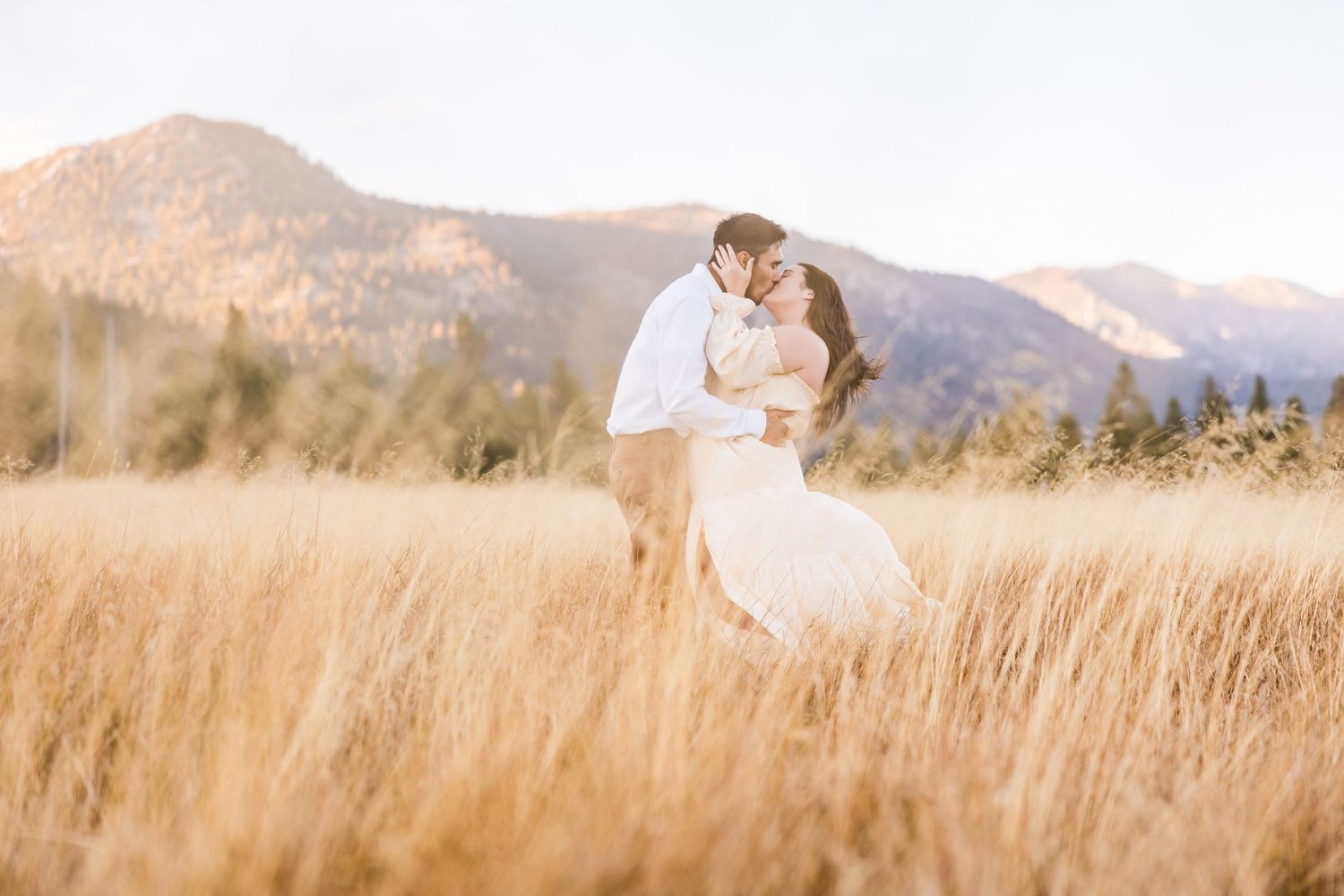 Couple holding hands in a green meadow with rolling hills, woman in white wedding dress, man in light blue shirt.